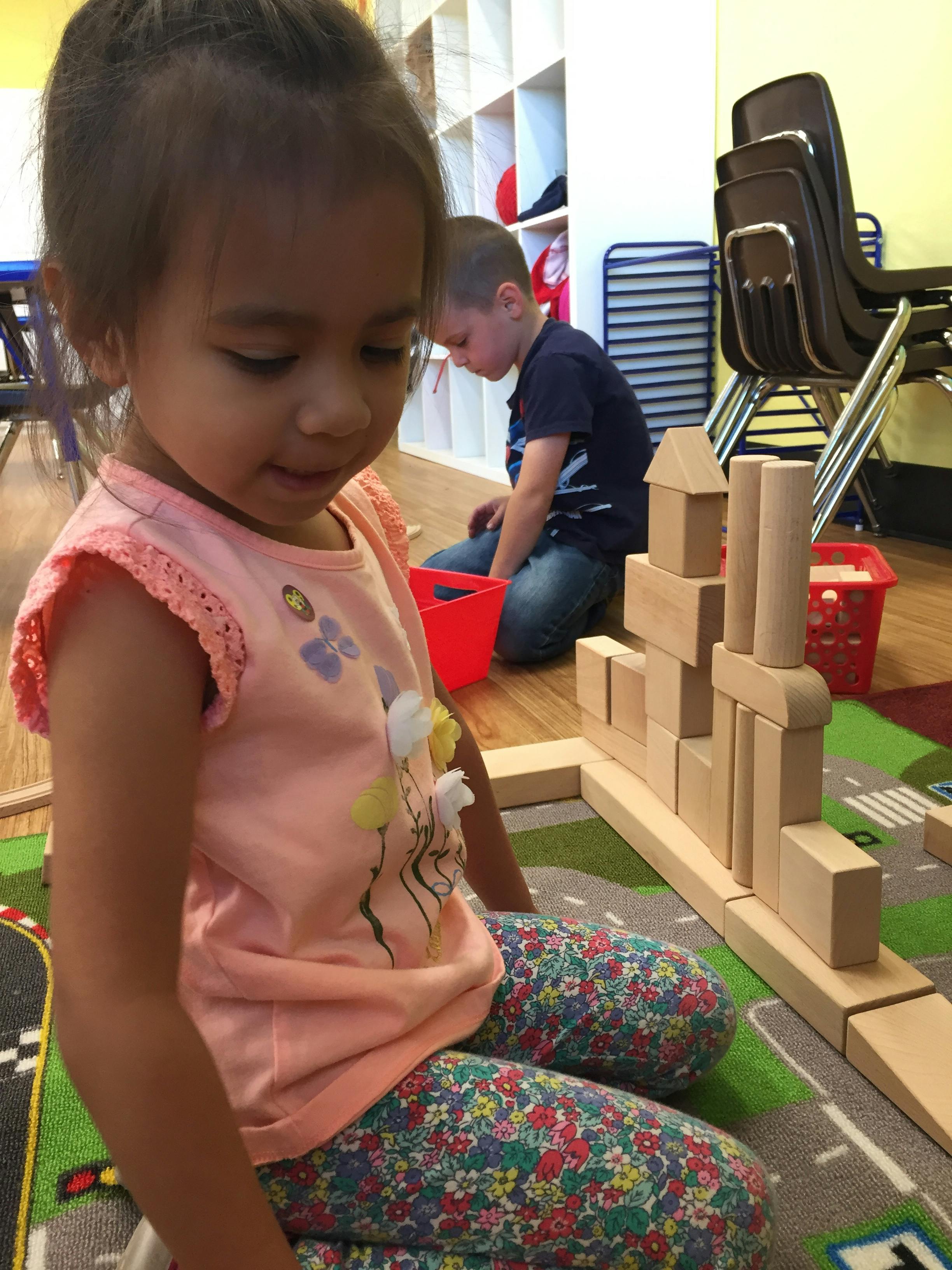 Child playing with wooden blocks on a rug, another child in background