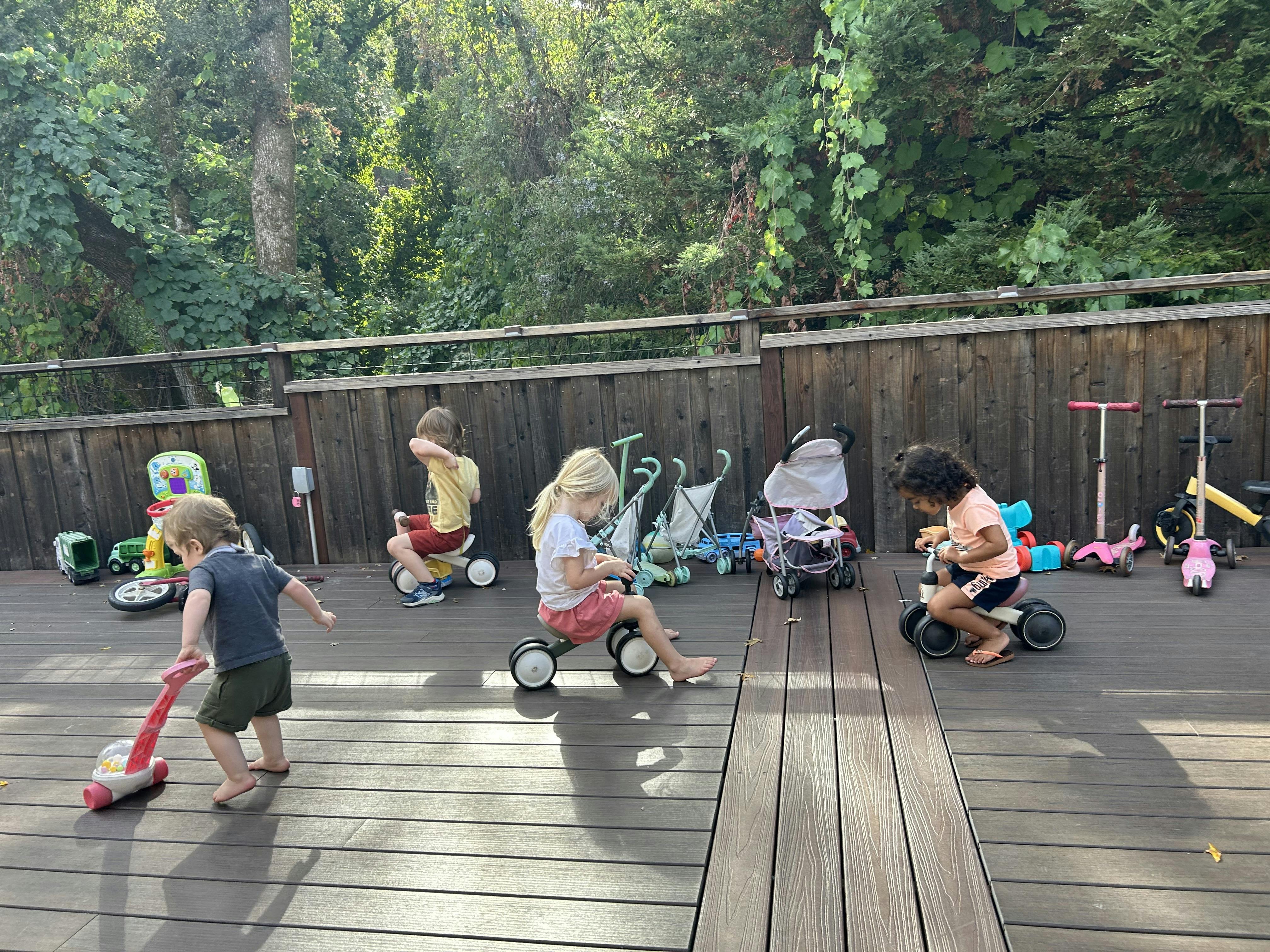 Children playing with ride-on toys and a toy vacuum on a deck