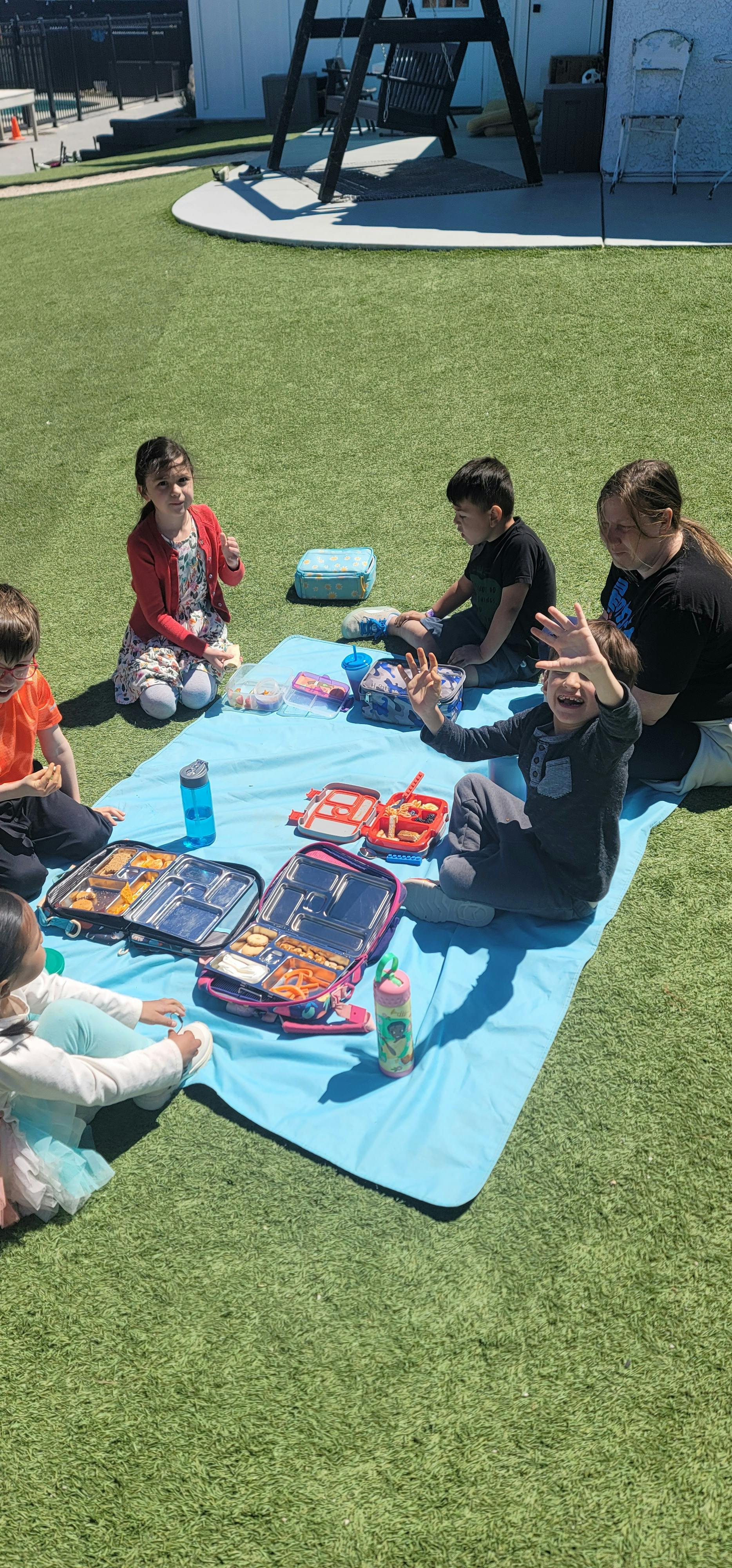Children and an adult sitting on a blue blanket on grass with open lunchboxes