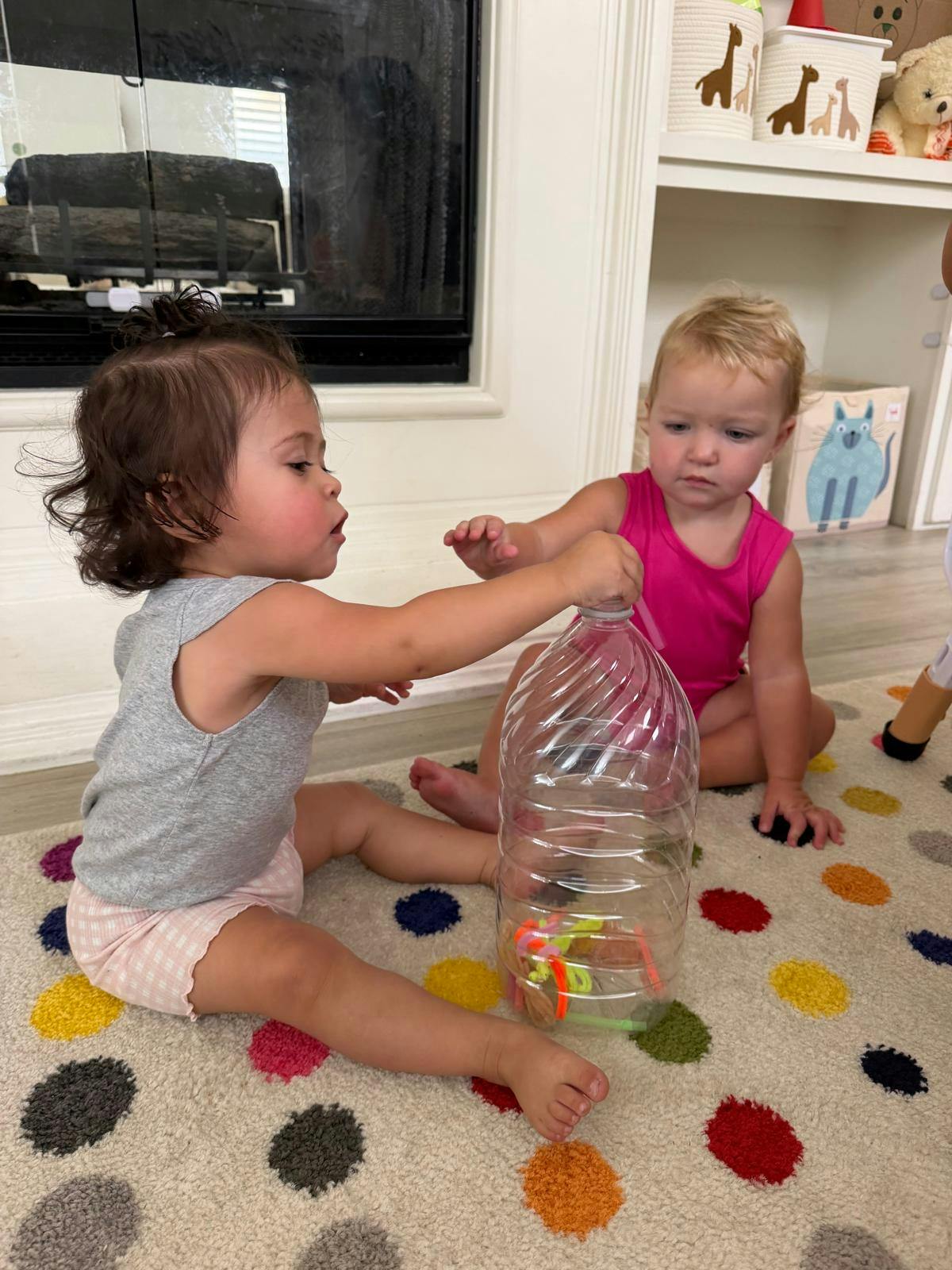 Two children sitting on a patterned rug playing with a plastic bottle