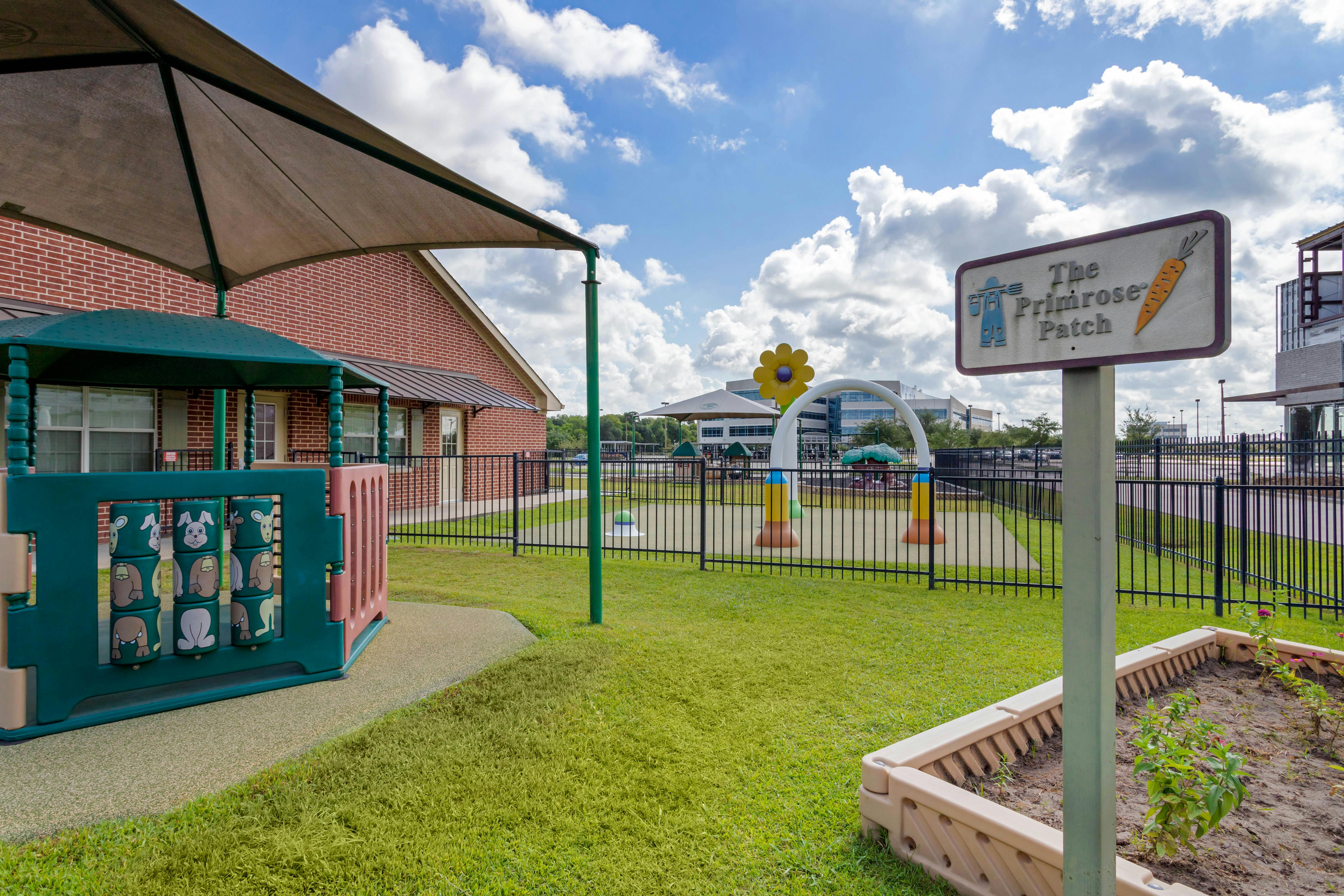 Playground area with shade canopy, brick building, and a sign for "The Primrose Patch"