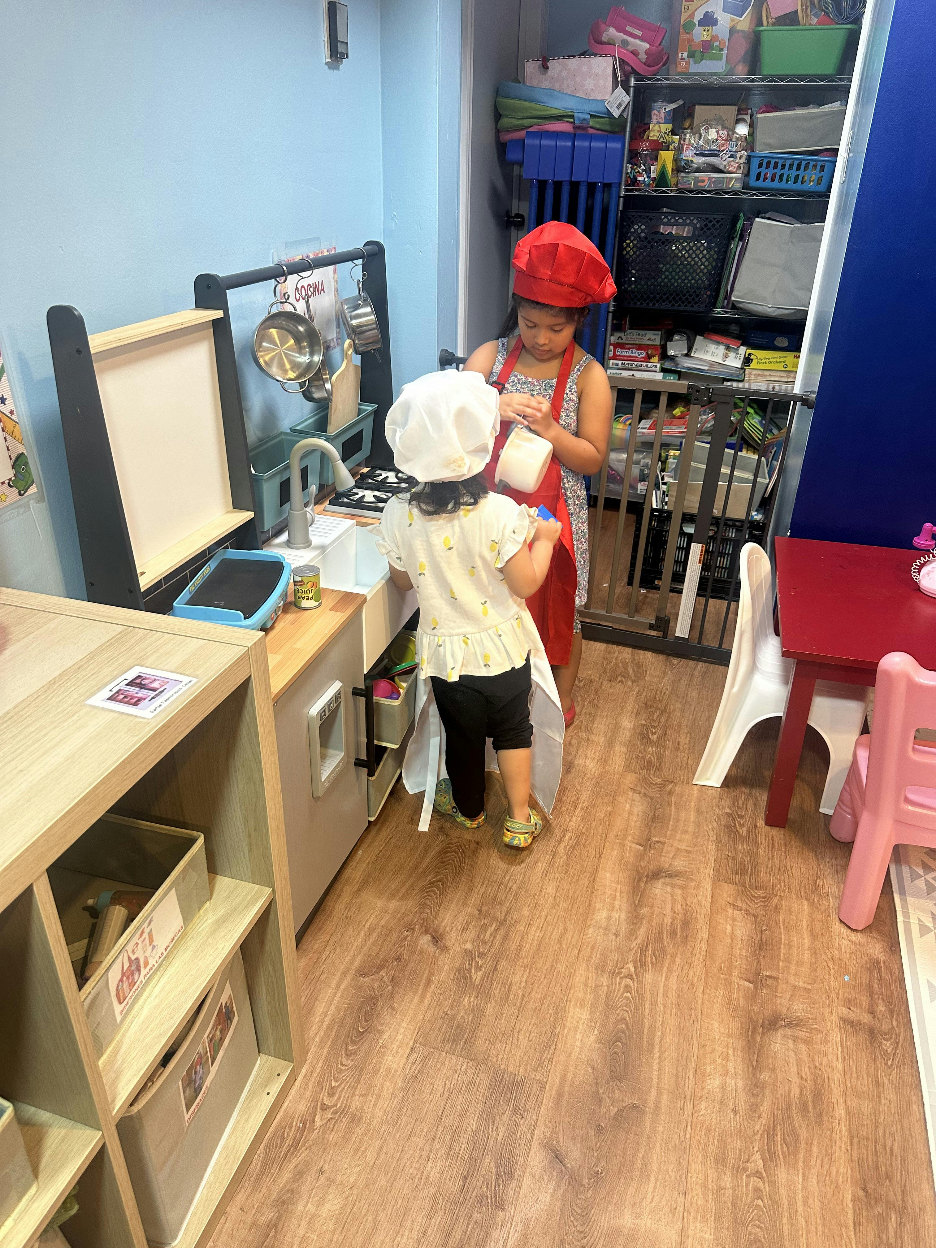 Two children playing in a toy kitchen, one wearing a red chef hat