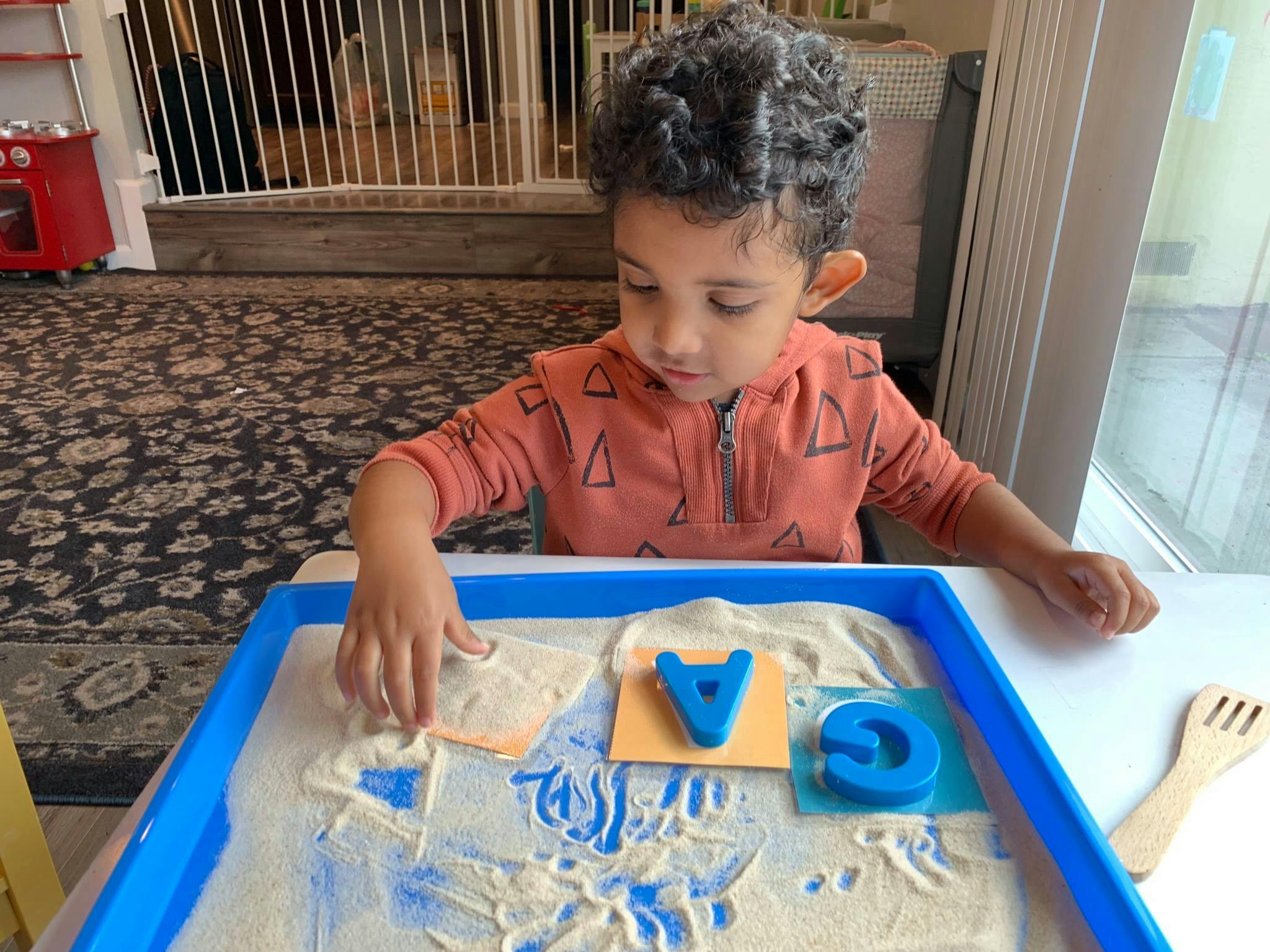 Child playing with sand and letter cutouts in a blue tray