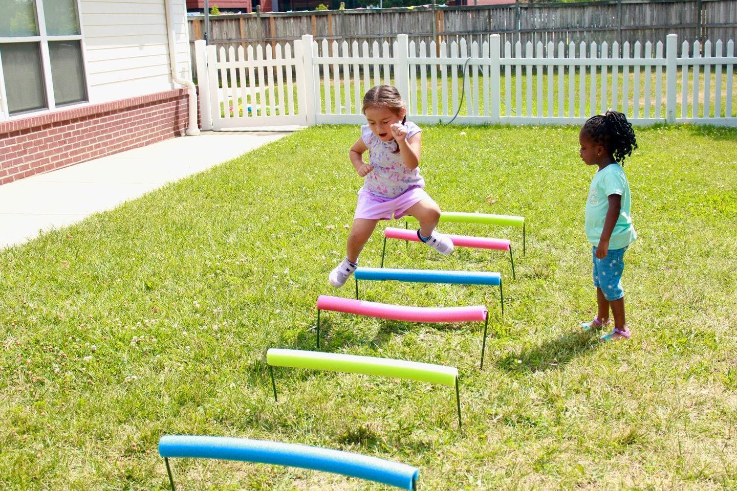 Child jumping over colorful hurdles in a grassy yard with another child watching