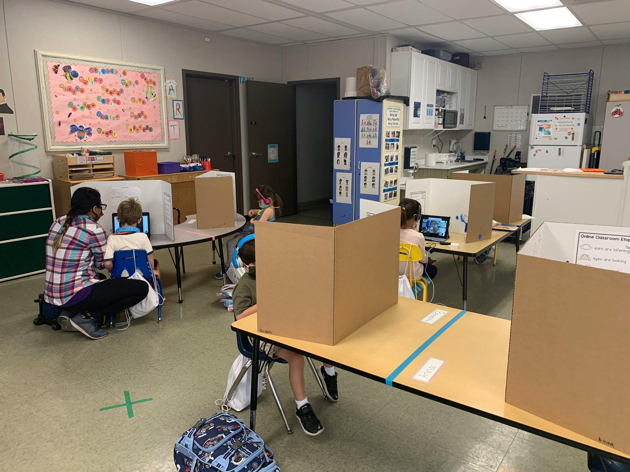 People and children working at desks with cardboard dividers in a classroom