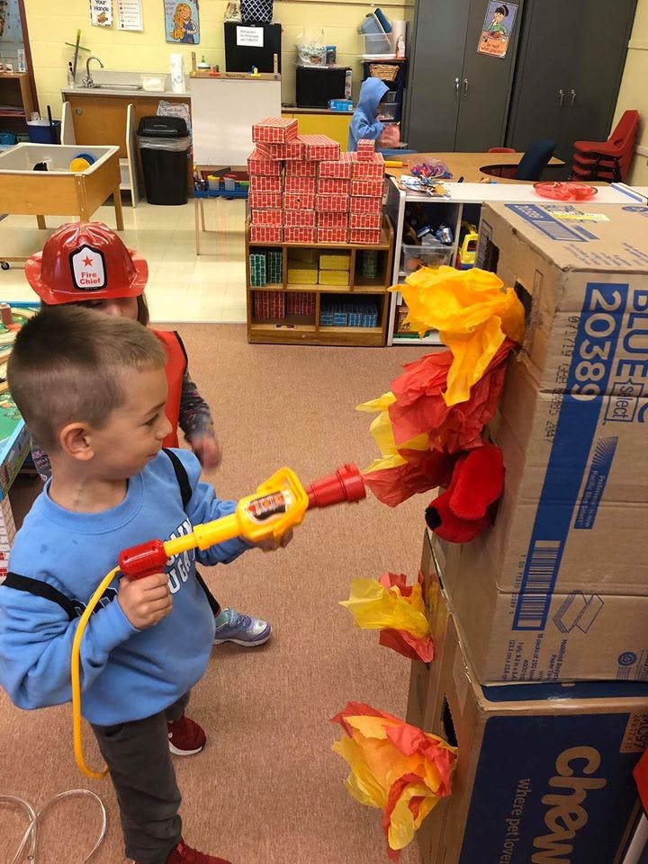 Child holding a toy fire hose pointed at a cardboard box with tissue paper flames