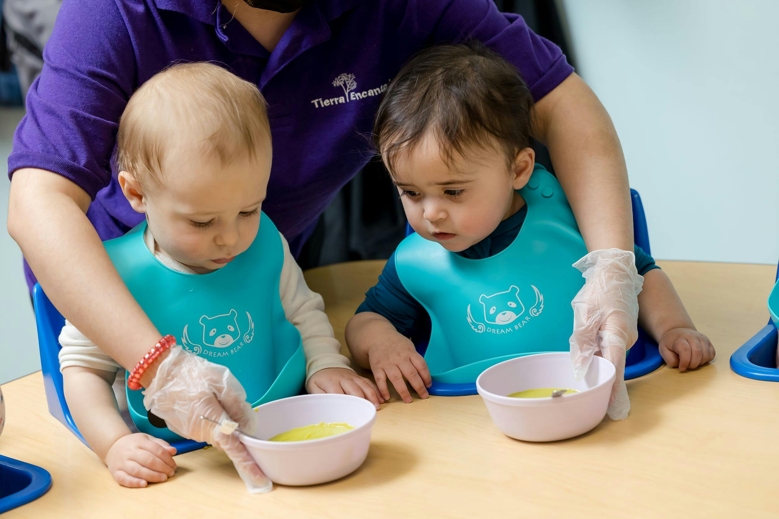 Person in gloves feeding two children in bibs at a table