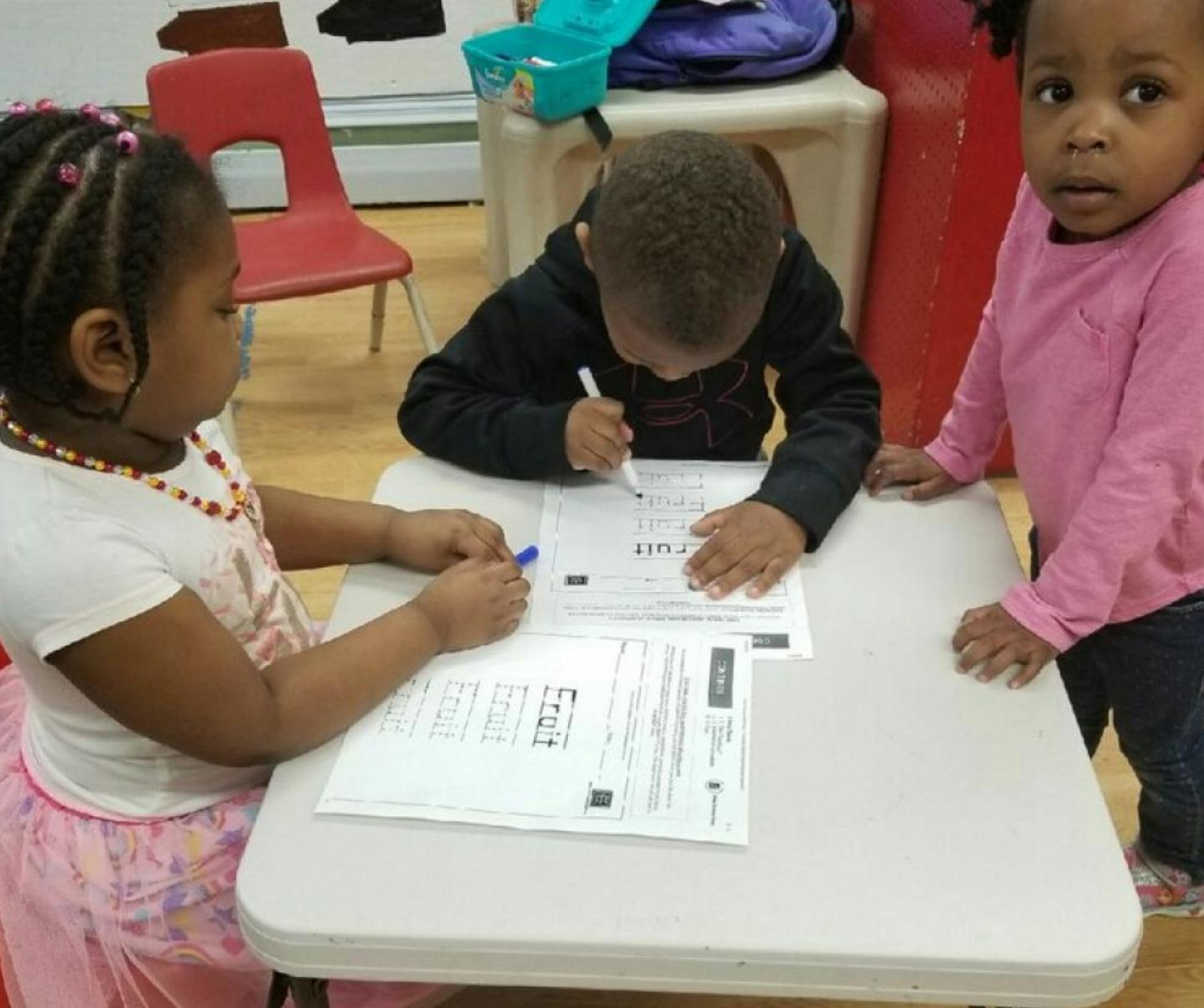 Three children at a table, two writing on papers, one standing