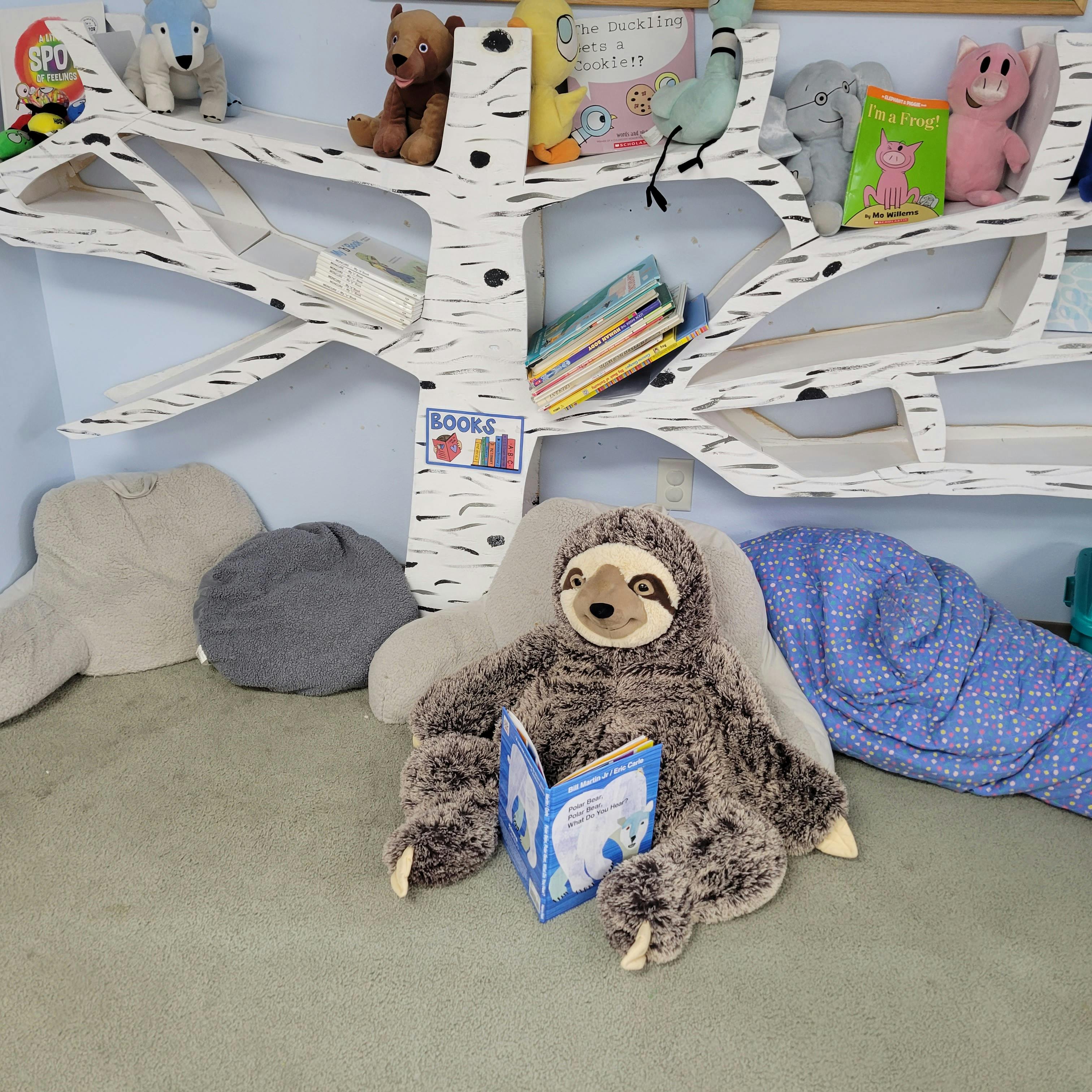 Sloth stuffed animal reading a book in front of a tree-shaped bookshelf