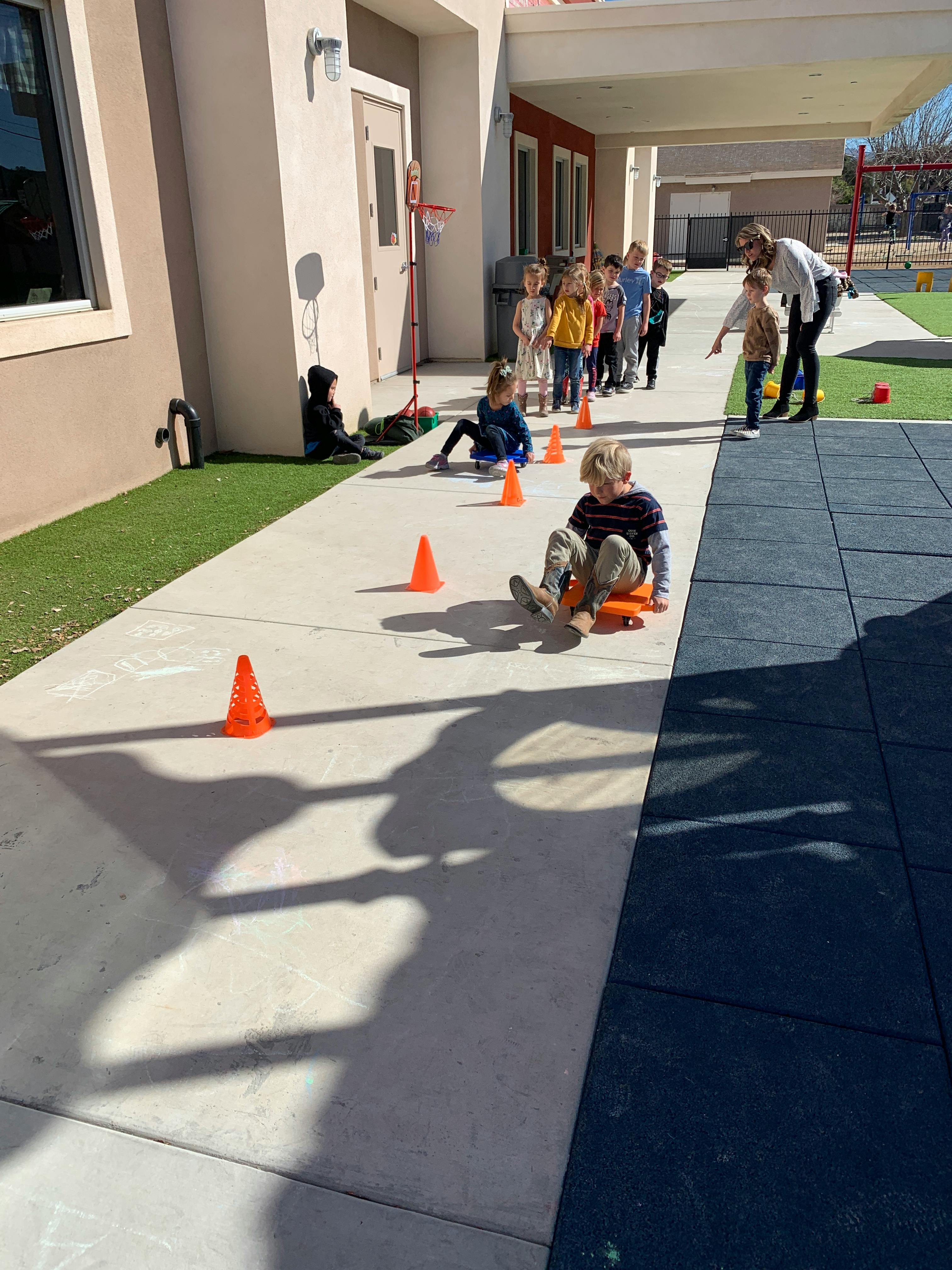 Children playing with scooters and cones on a sidewalk with an adult supervising