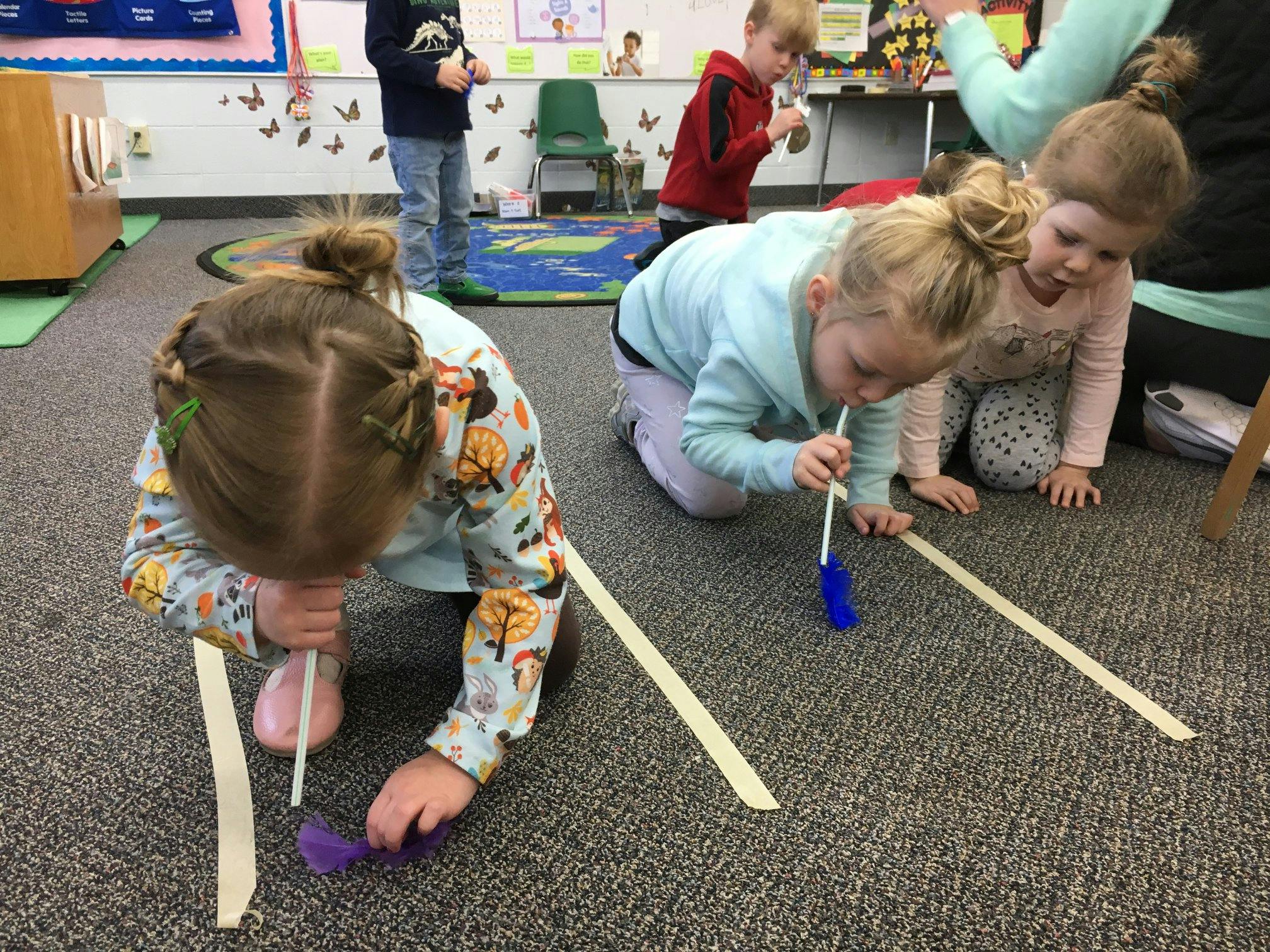 Children on floor blowing objects with straws in a classroom