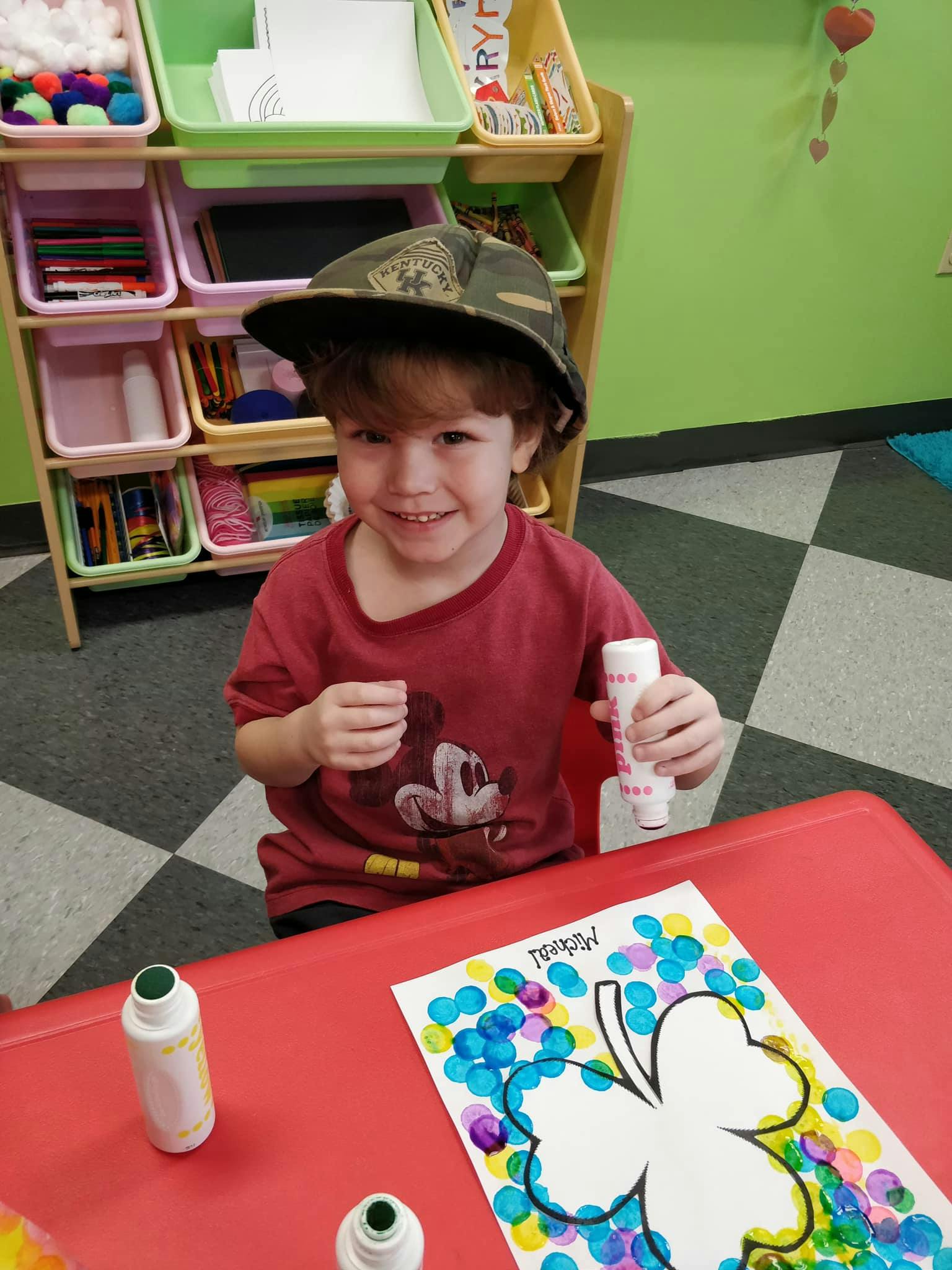 Child in a hat holding a marker, sitting at a table with a shamrock craft