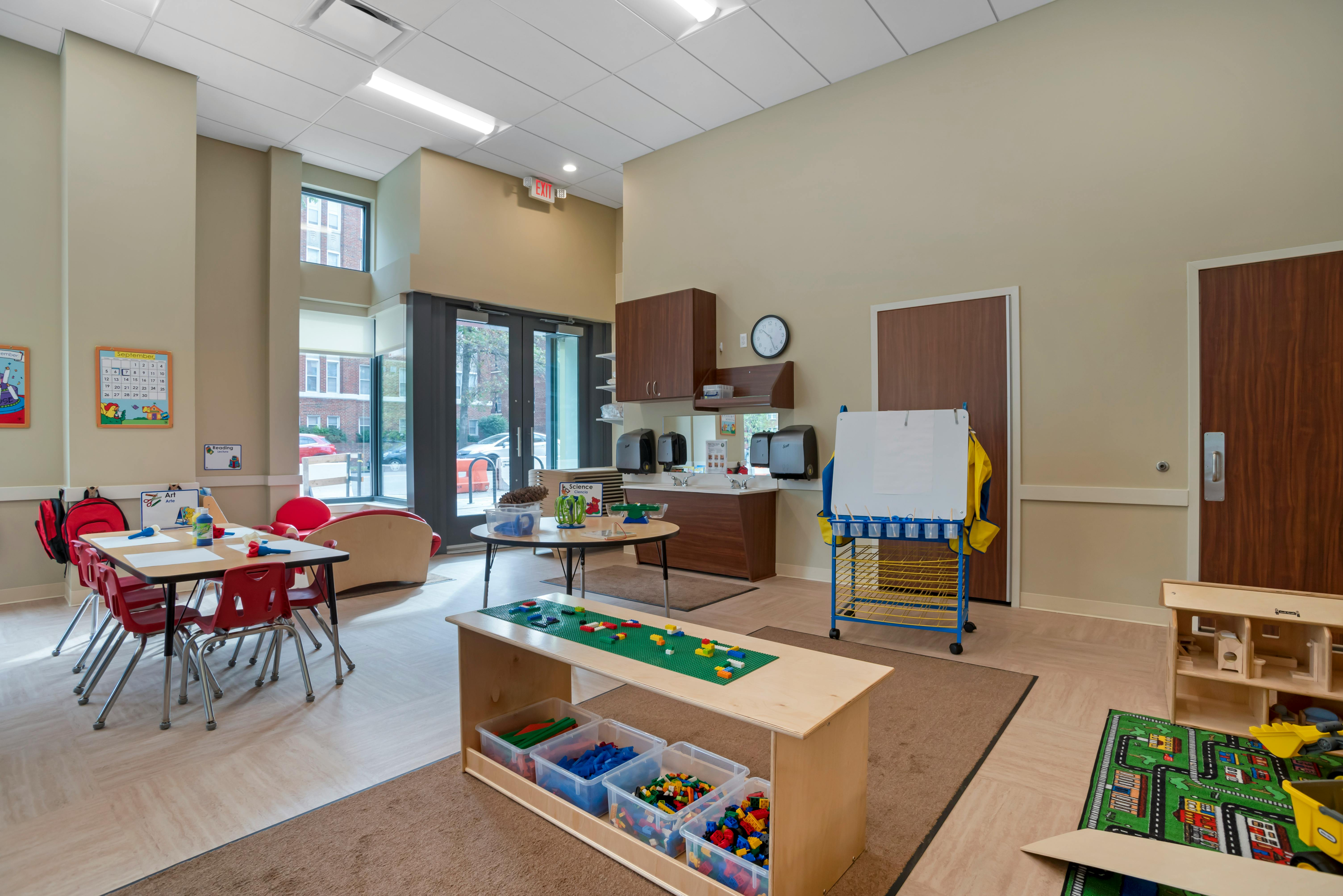 Classroom with small red chairs at tables, a sink counter, and play area with blocks