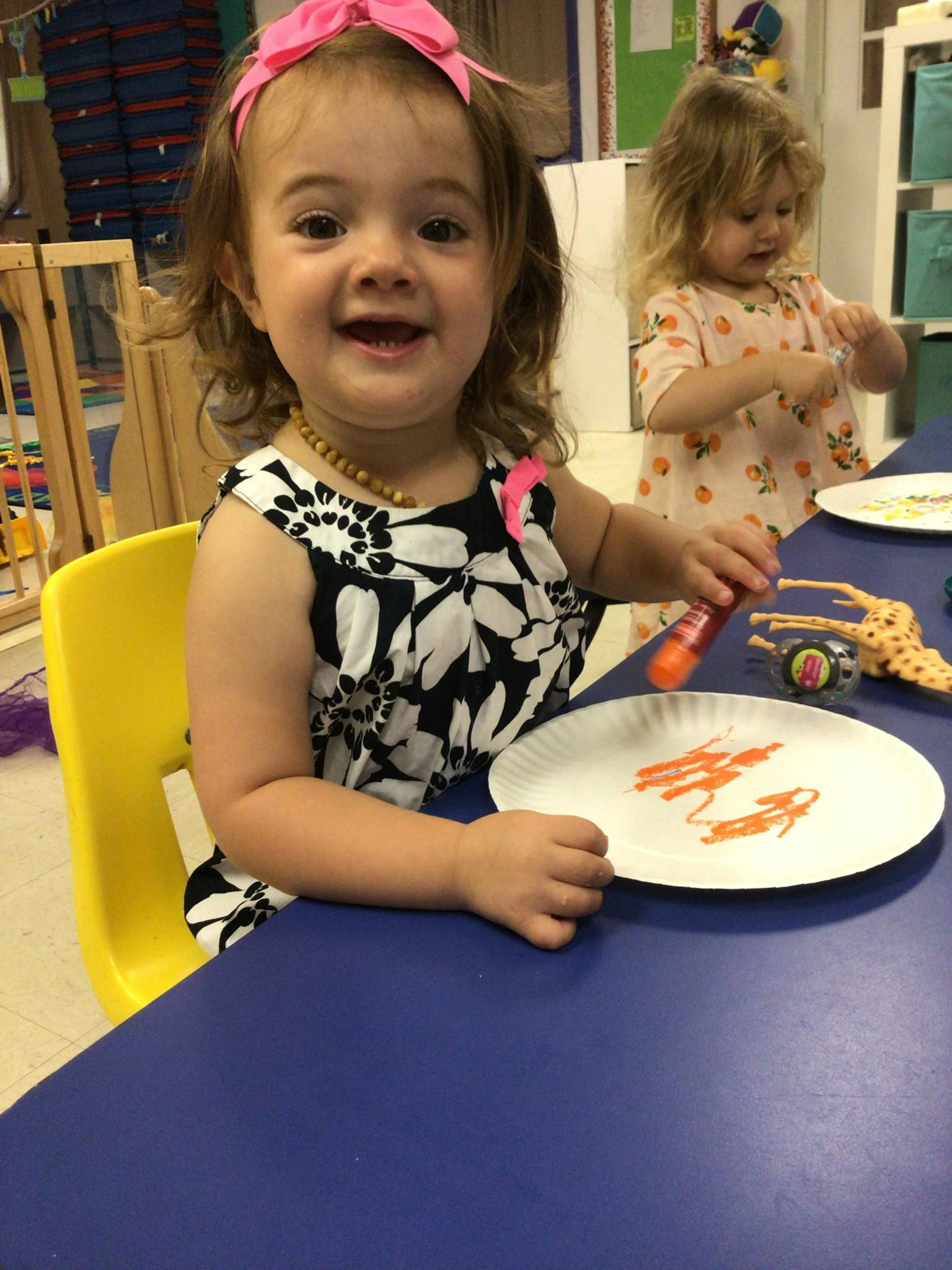 Child sitting at a table with a plate and a squeeze bottle, another child in background