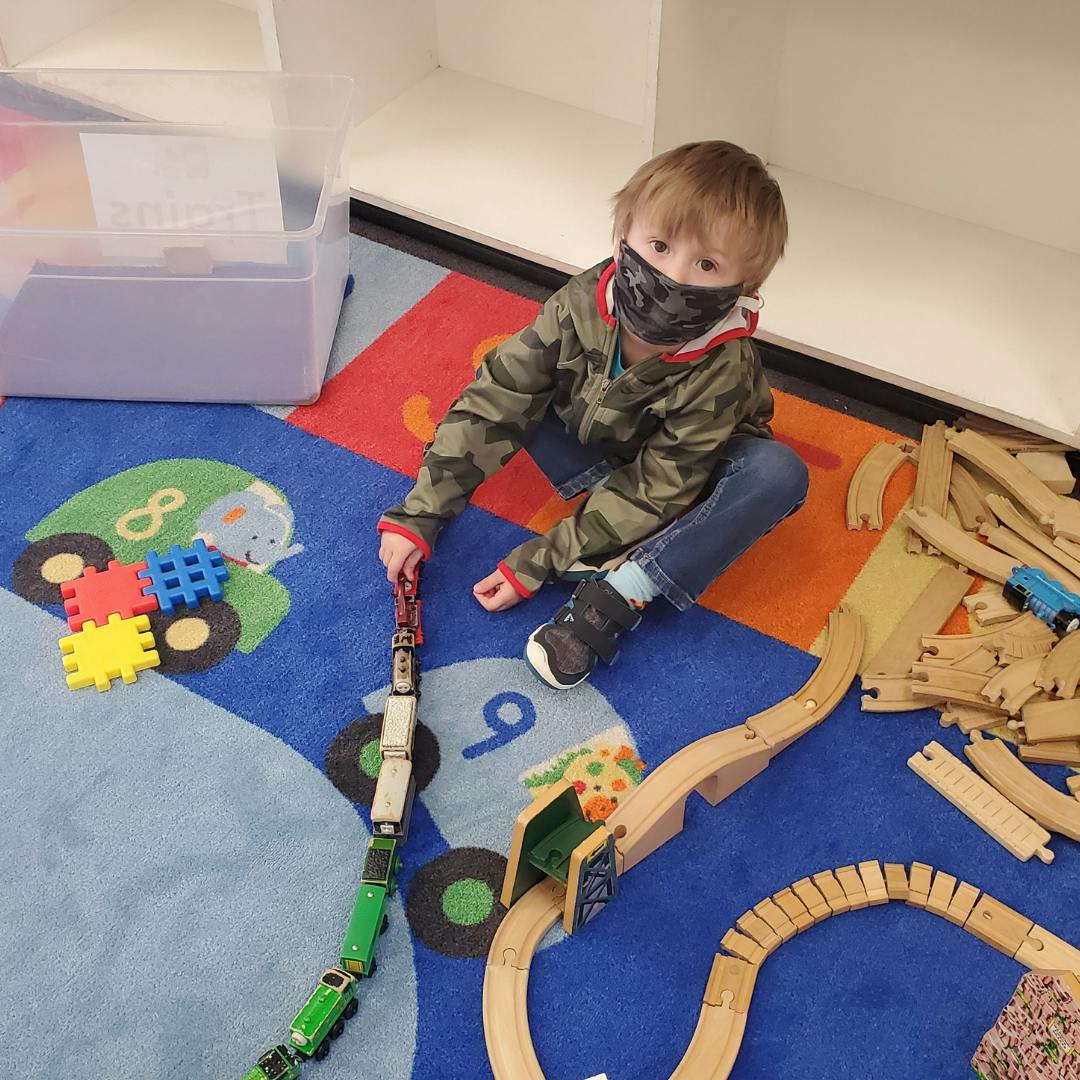 Child in a mask playing with a toy train and wooden tracks on a colorful rug