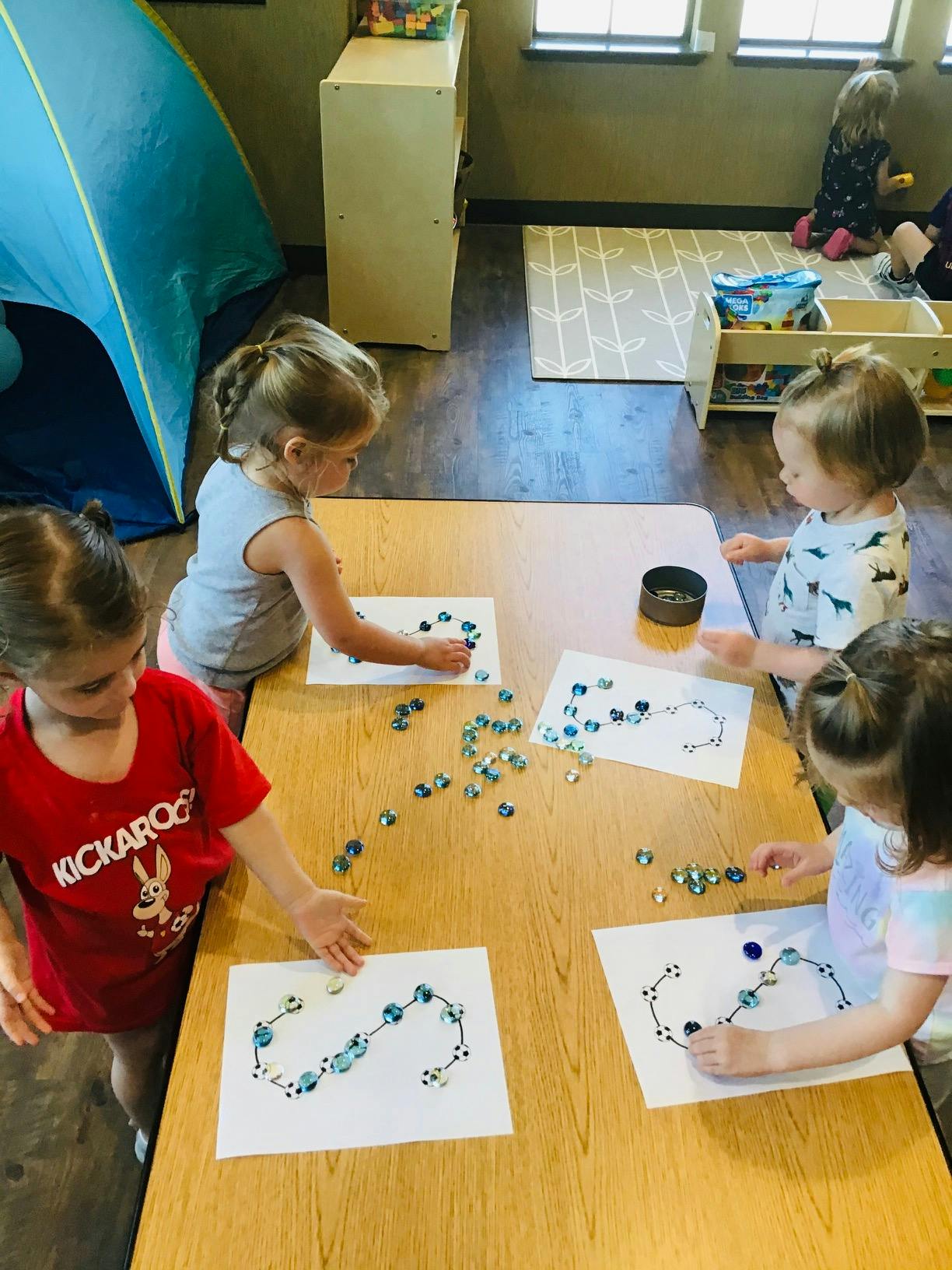 Four children placing small blue and clear beads on papers at a table