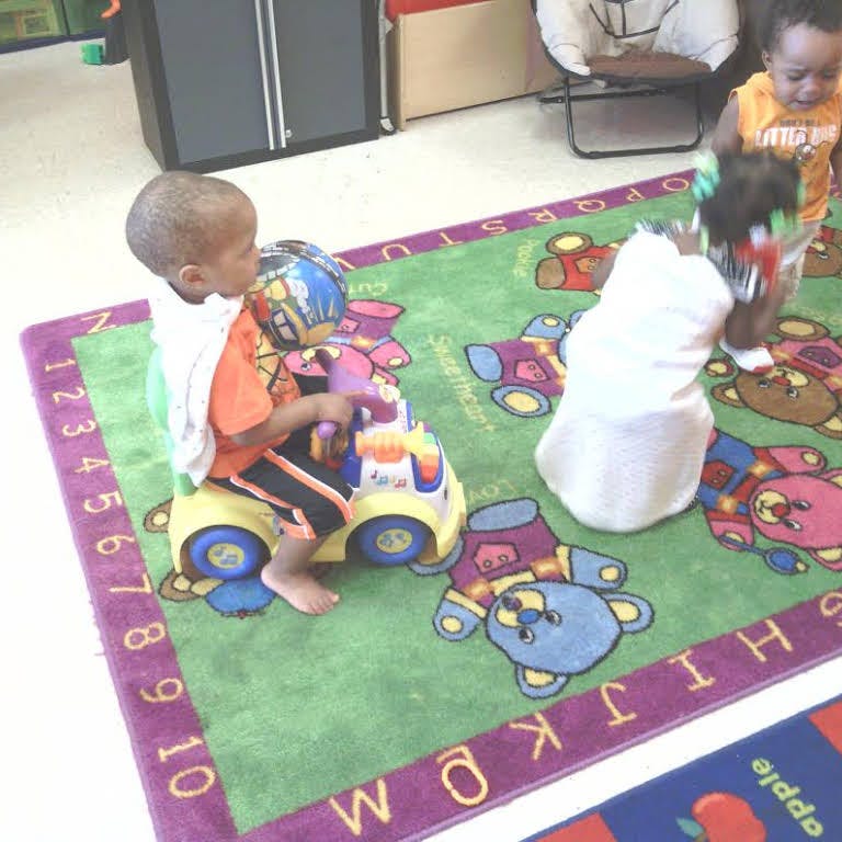 Child sitting on a toy car on a rug with other children nearby