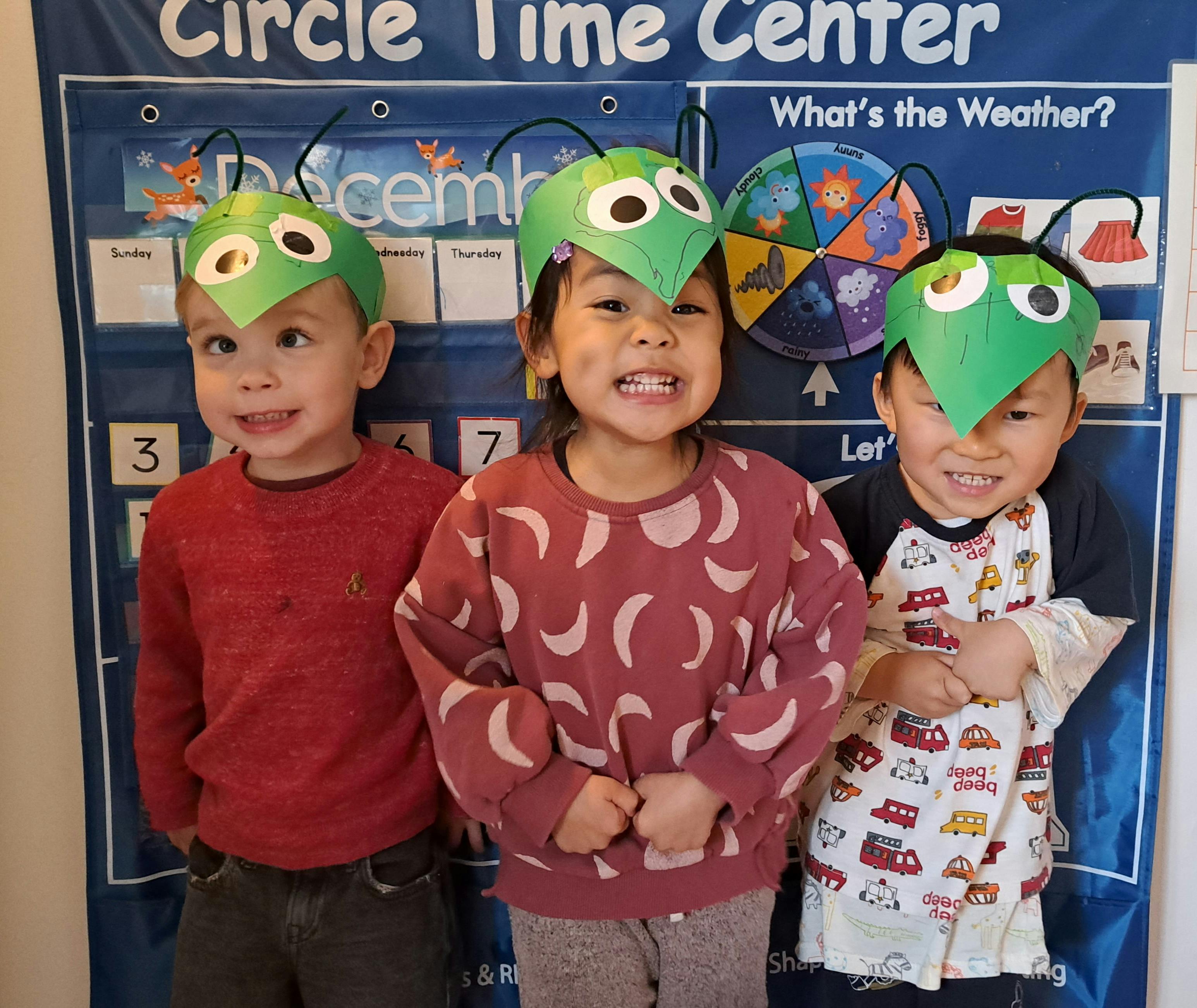 Three children wearing green bug headbands in front of a calendar