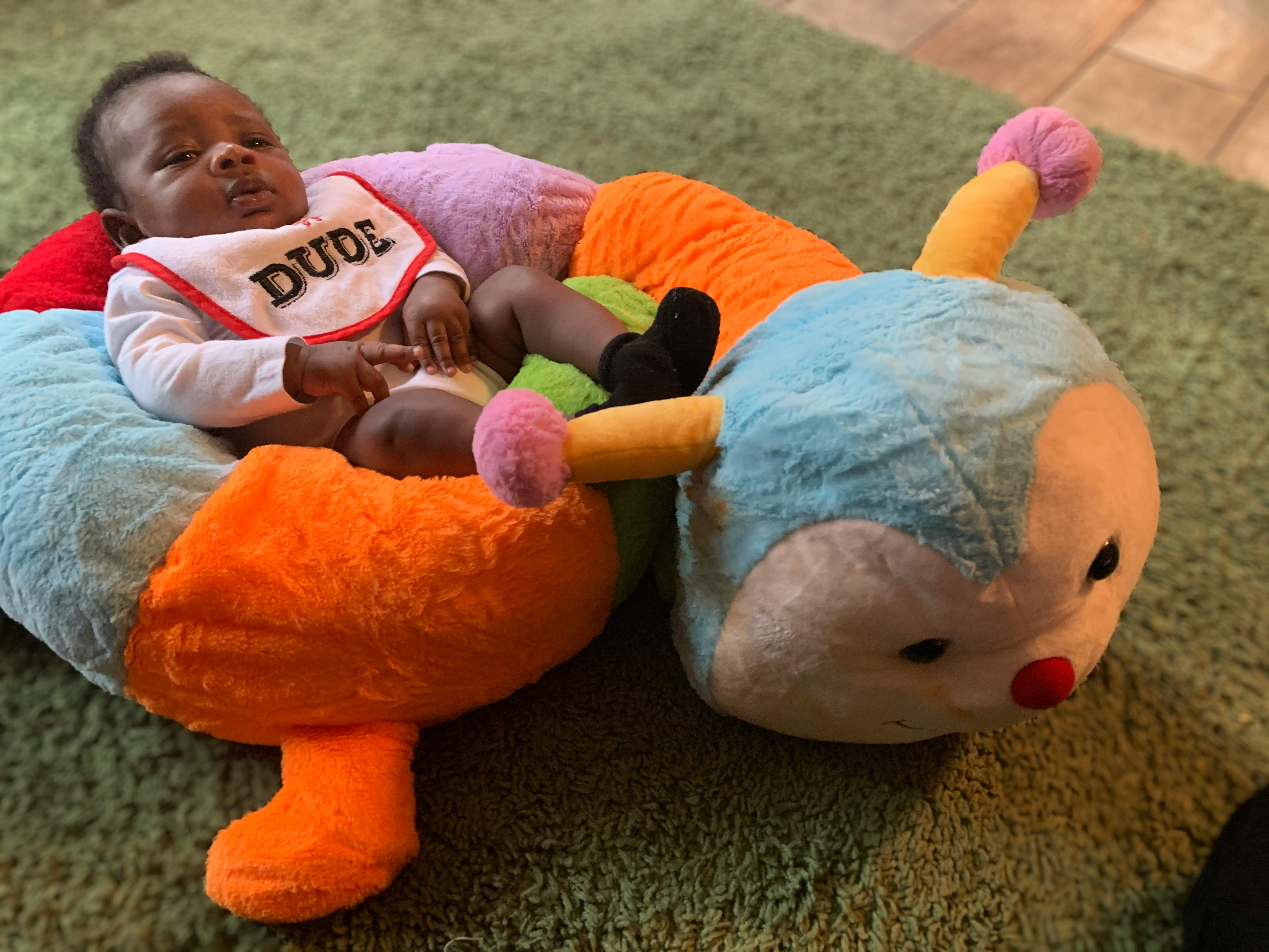Child wearing a bib with "DUDE" on it, sitting in a caterpillar-shaped plush toy