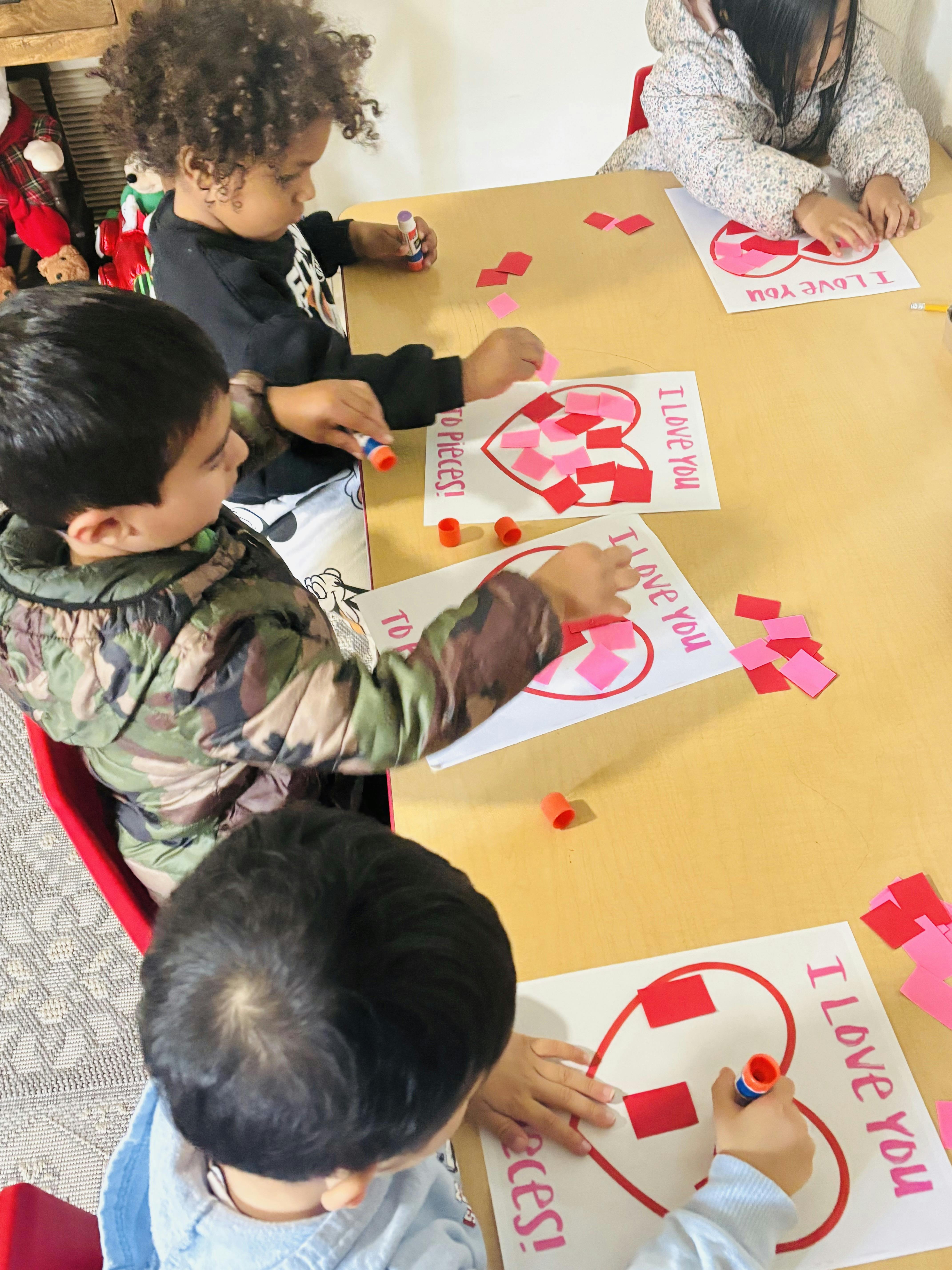 Children gluing red and pink squares onto heart shapes that say "I Love You"