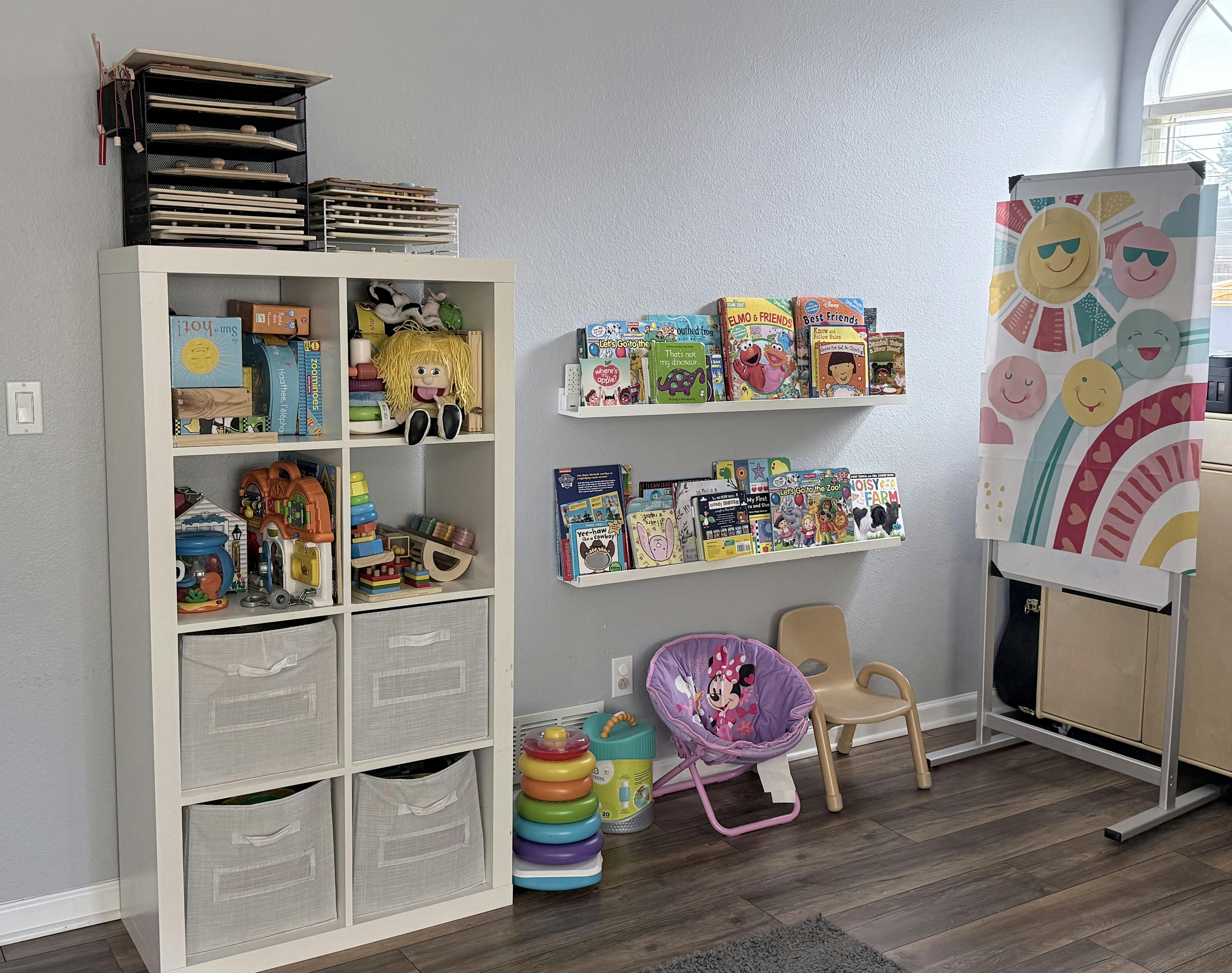 Children's room with a white shelving unit holding toys and bins, wall-mounted book shelves with shelves, and a child's easel