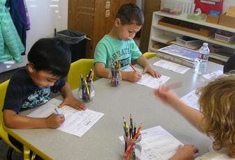 Three children writing at a table in a classroom