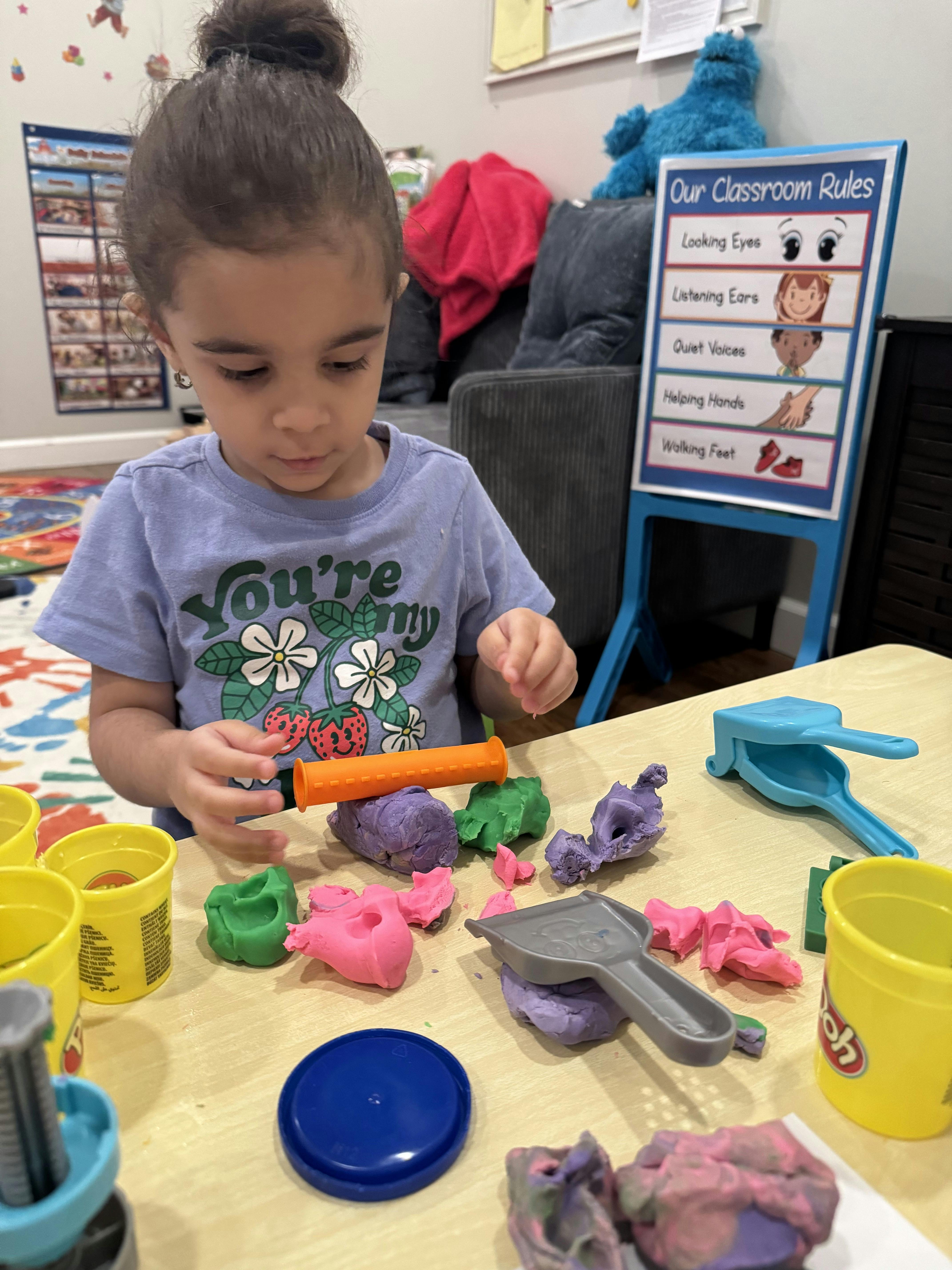 Child playing with Play-Doh on a table, with a \"Classroom Rules\" chart in the background