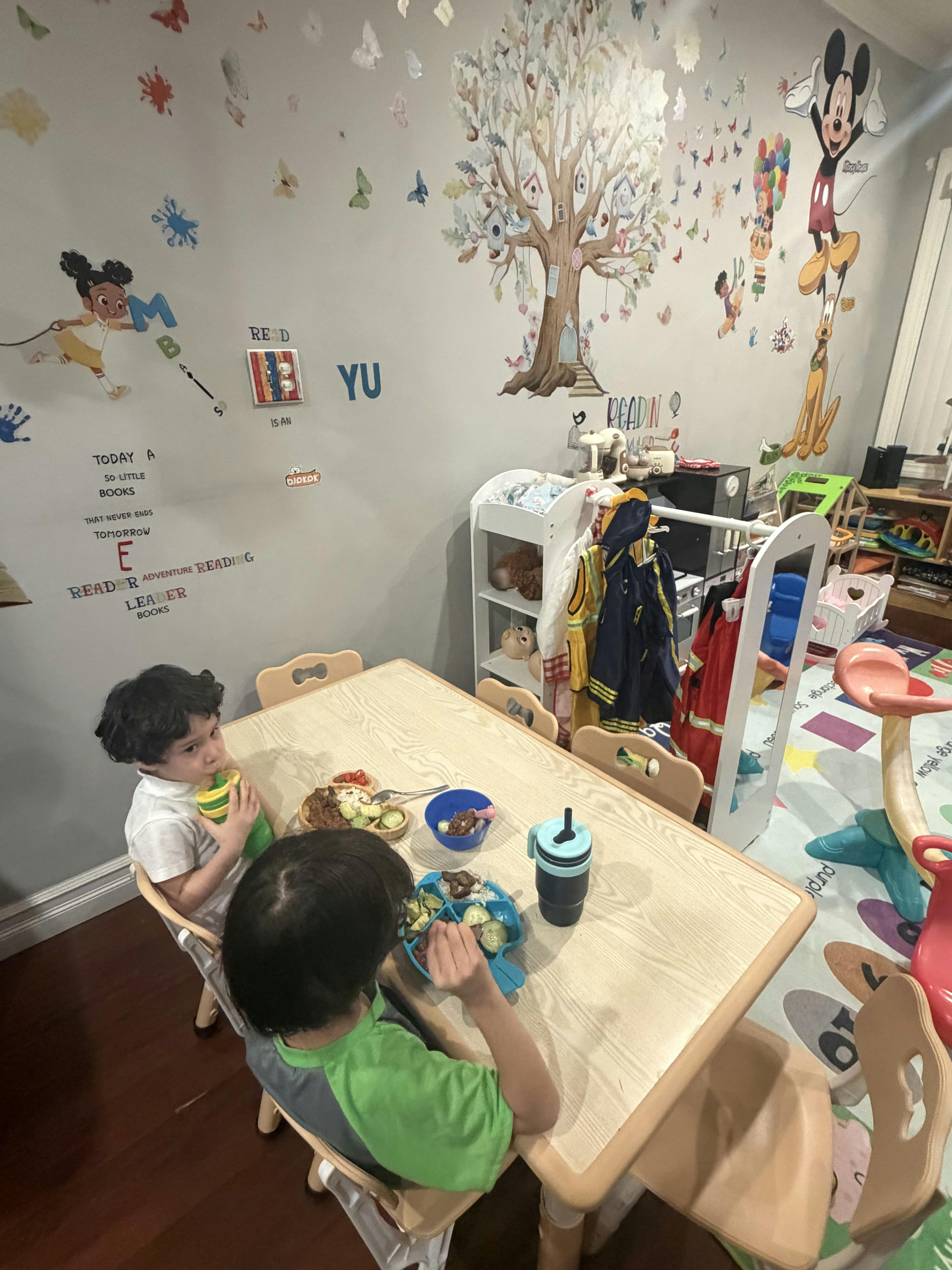 Two children seated at a table eating, one drinking from a green cup