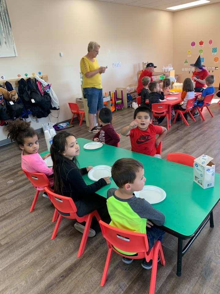 Children sitting at tables in a classroom with an adult standing nearby