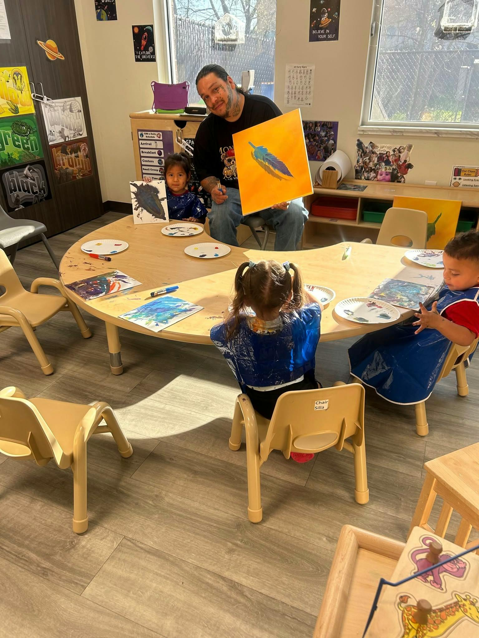 Adult holding up a painting to children seated at a table in a classroom