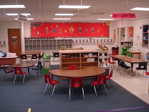 Empty classroom with small tables and chairs, a red bulletin board, and storage cubbies