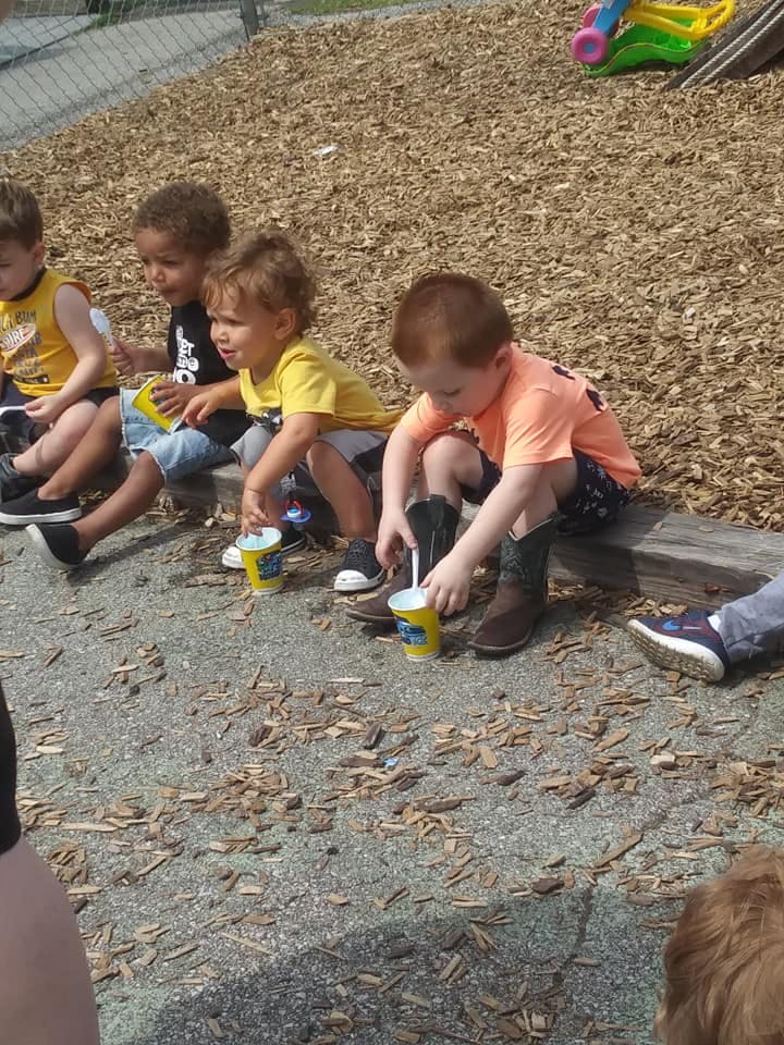 Children sitting on a wooden beam, one child stirring a cup with a straw