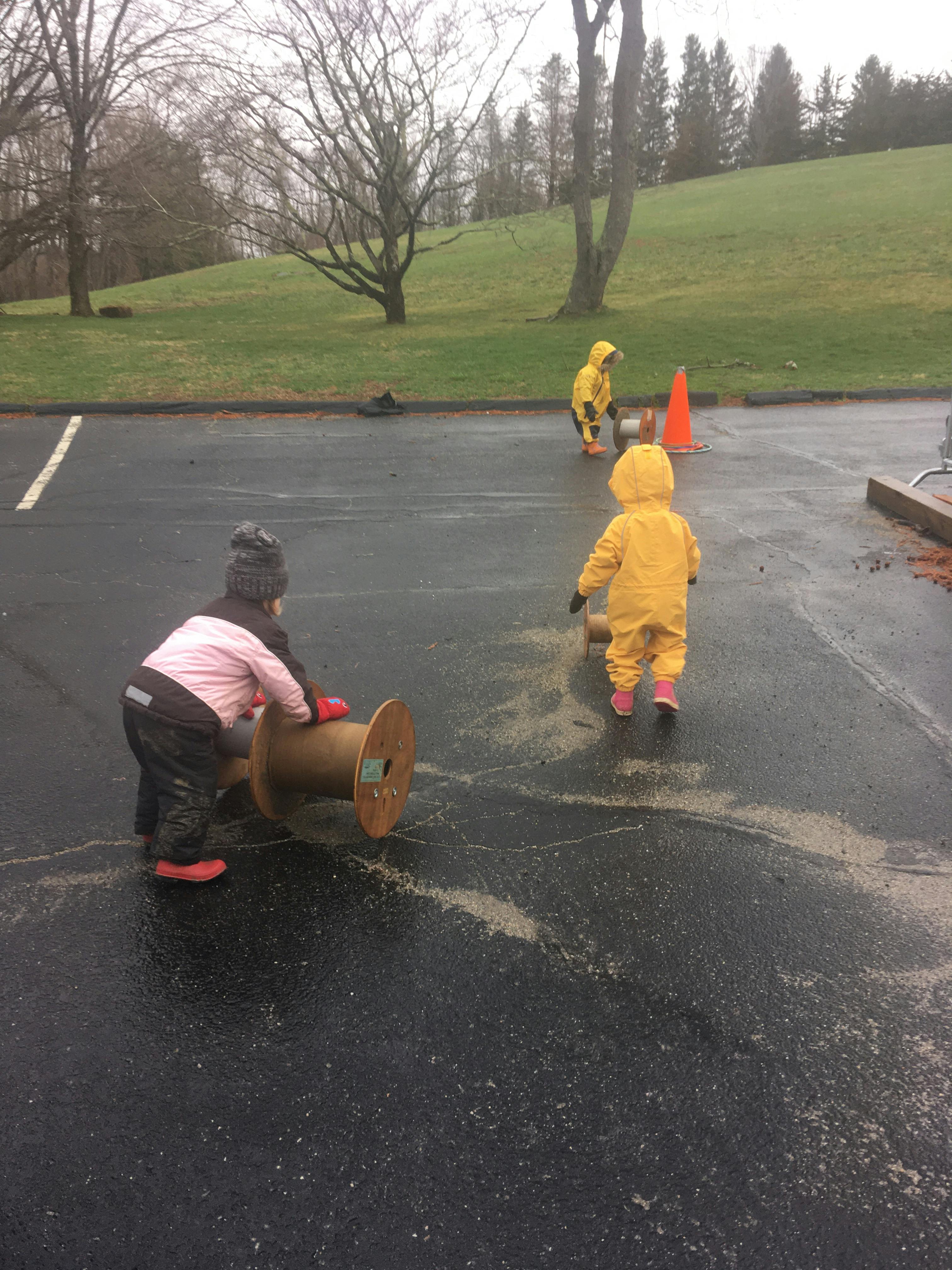 Child pushing a large wooden spool on pavement, two other children nearby
