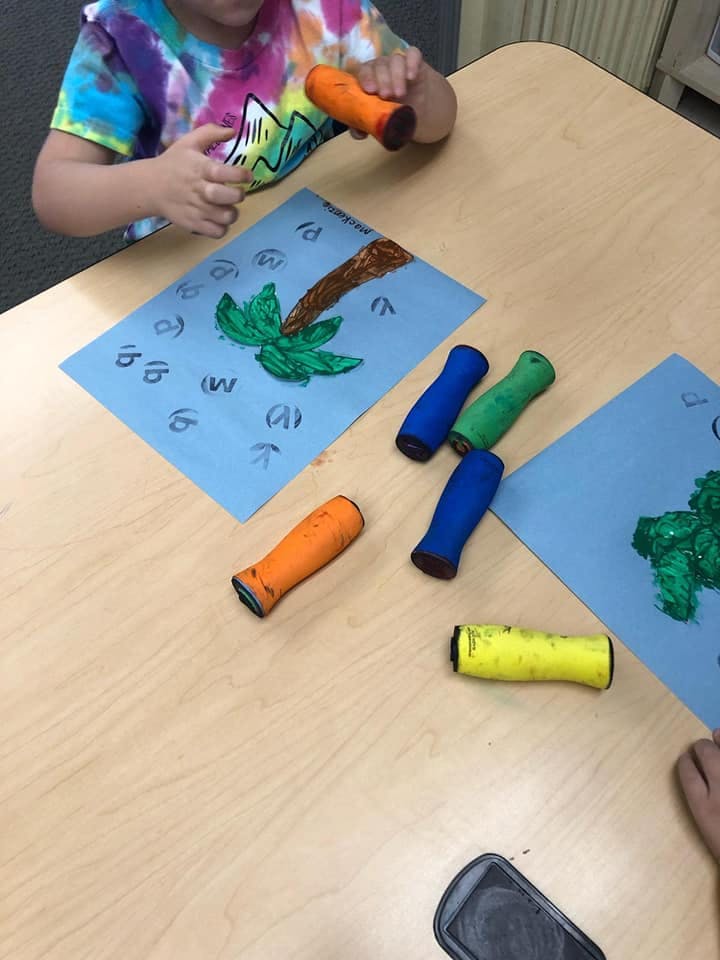 Child holding an orange paint roller over a blue paper with a painted palm tree