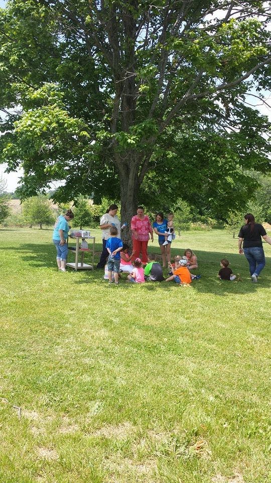People and children gathered under a large tree in a grassy field