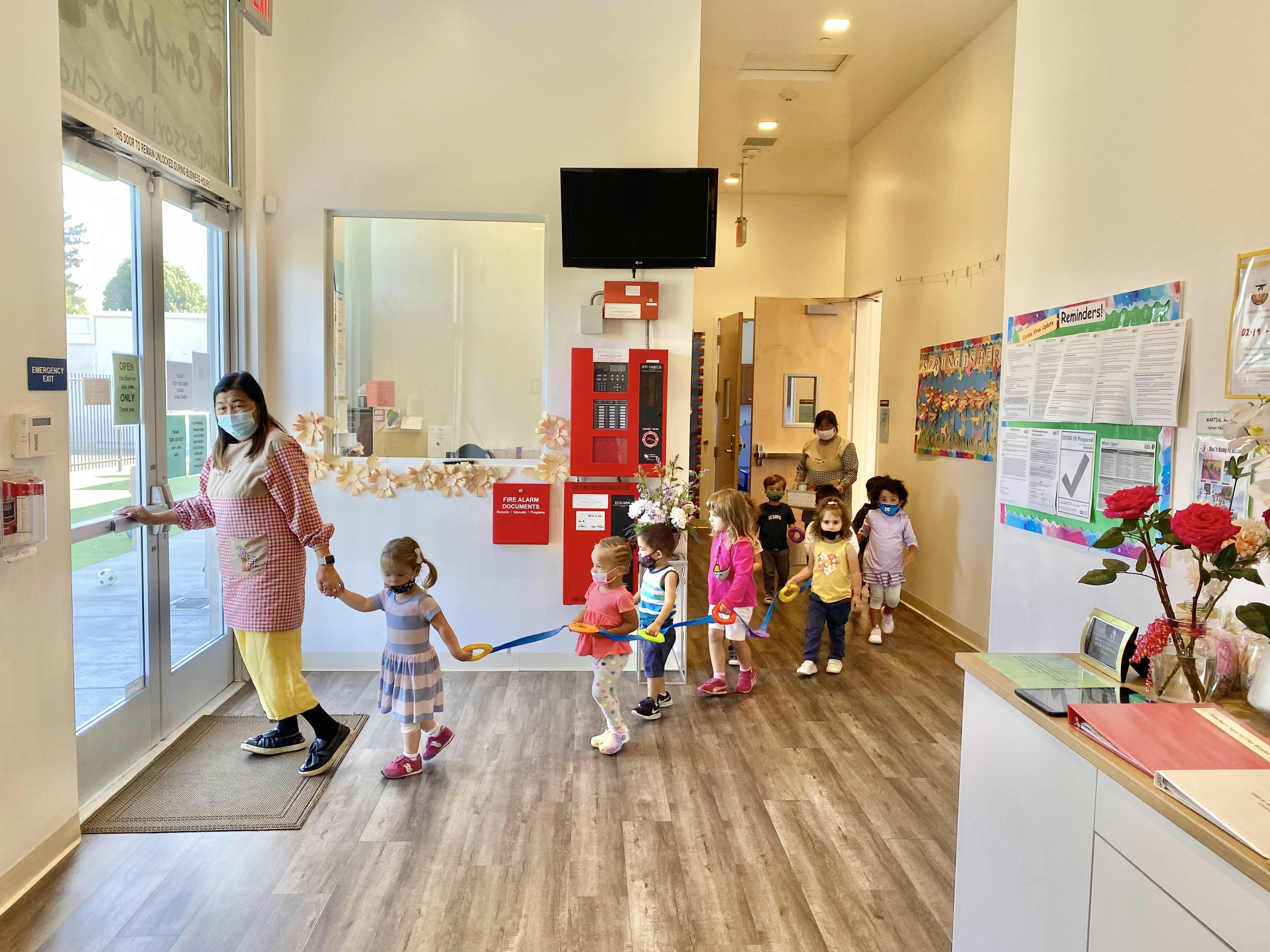 Person in mask opening door for line of children holding a walking rope