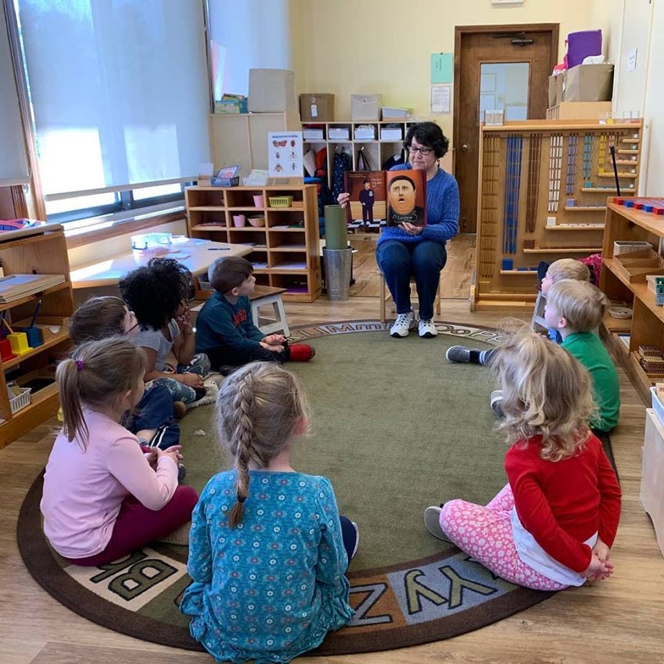 Person reading a book to a group of children sitting on a rug in a classroom