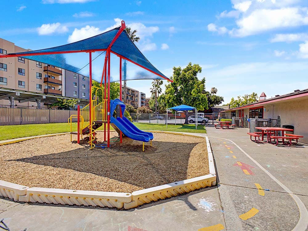 A playground with a blue and red structure and slides covered by a shade sail