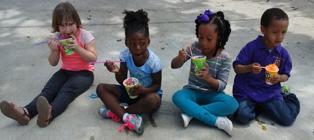 Four children sitting on the ground eating from cups with spoons and without spoons