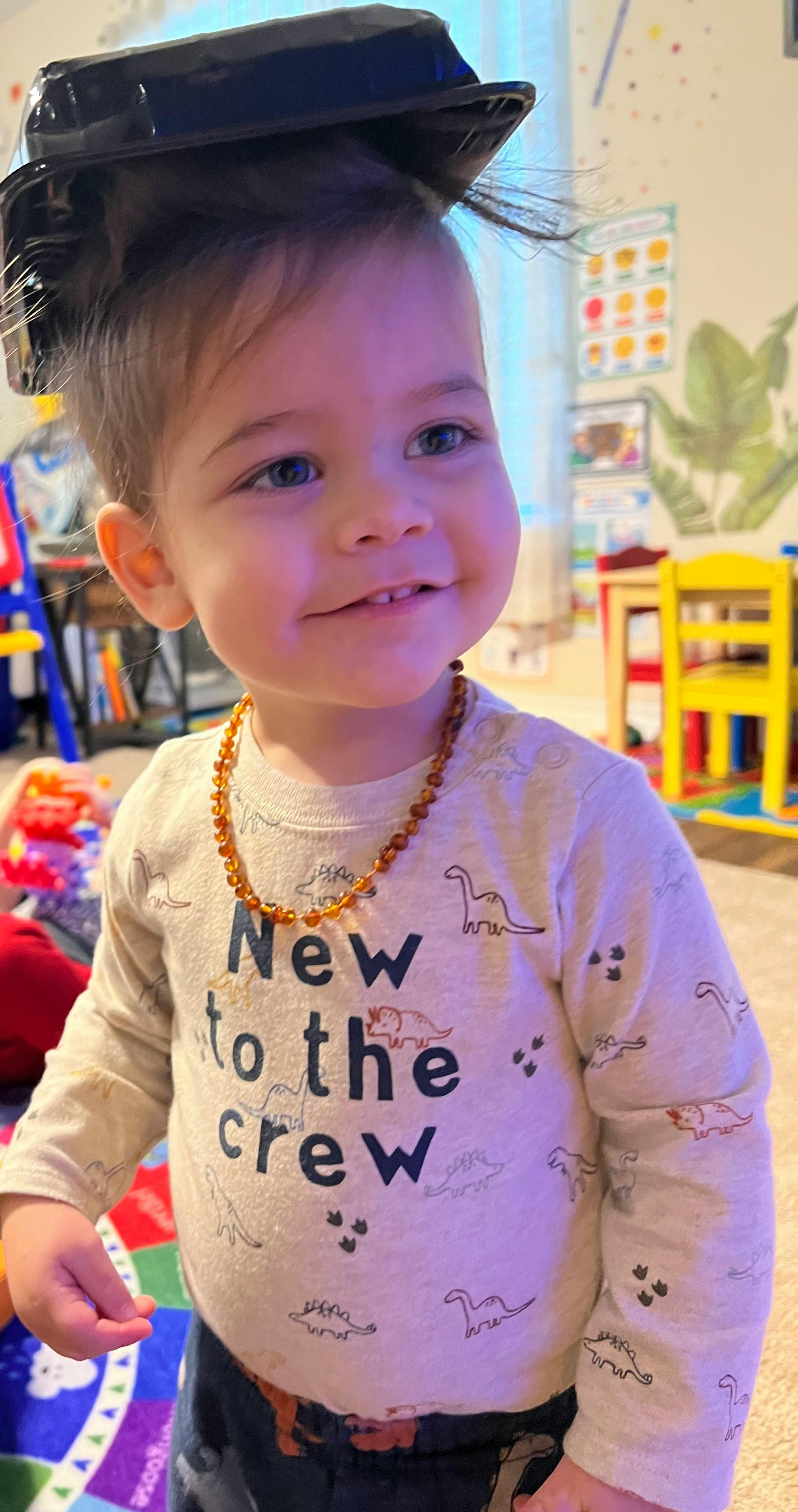 Child with a black container on their head, wearing a shirt that says "New to the crew"