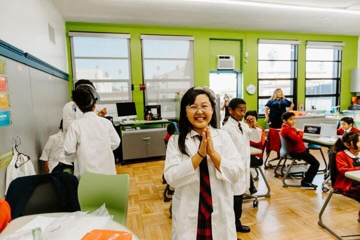 Child in a lab coat smiling in a classroom with other children and an adult