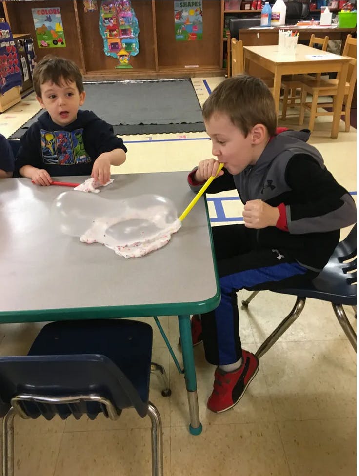 Child blowing into a straw to make a bubble on a table, another child watching