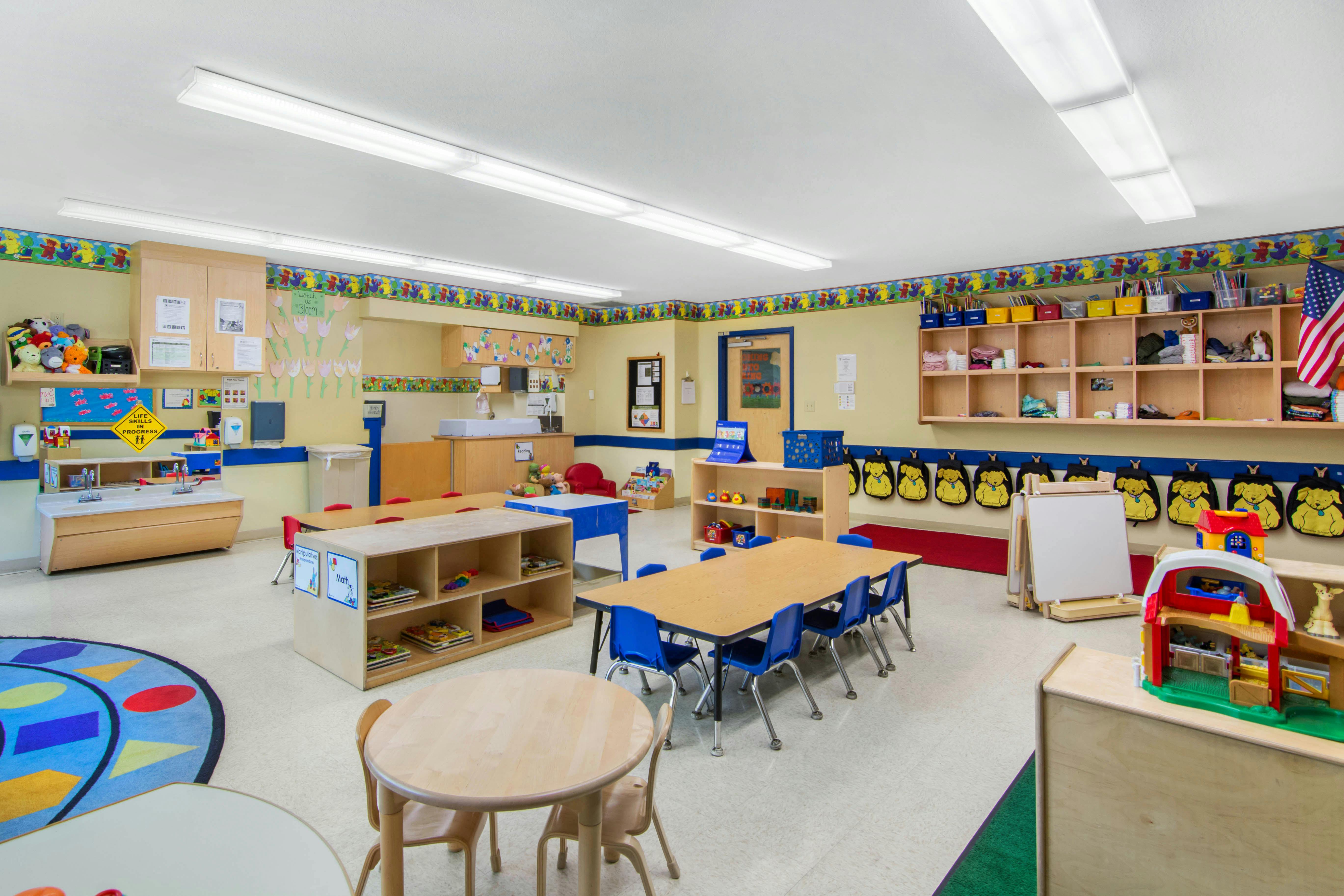 Classroom with child-sized tables and chairs, shelves, and a rug with shapes