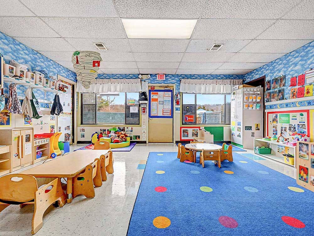 Classroom with child-sized tables and chairs, a blue rug with colored dots, and toys