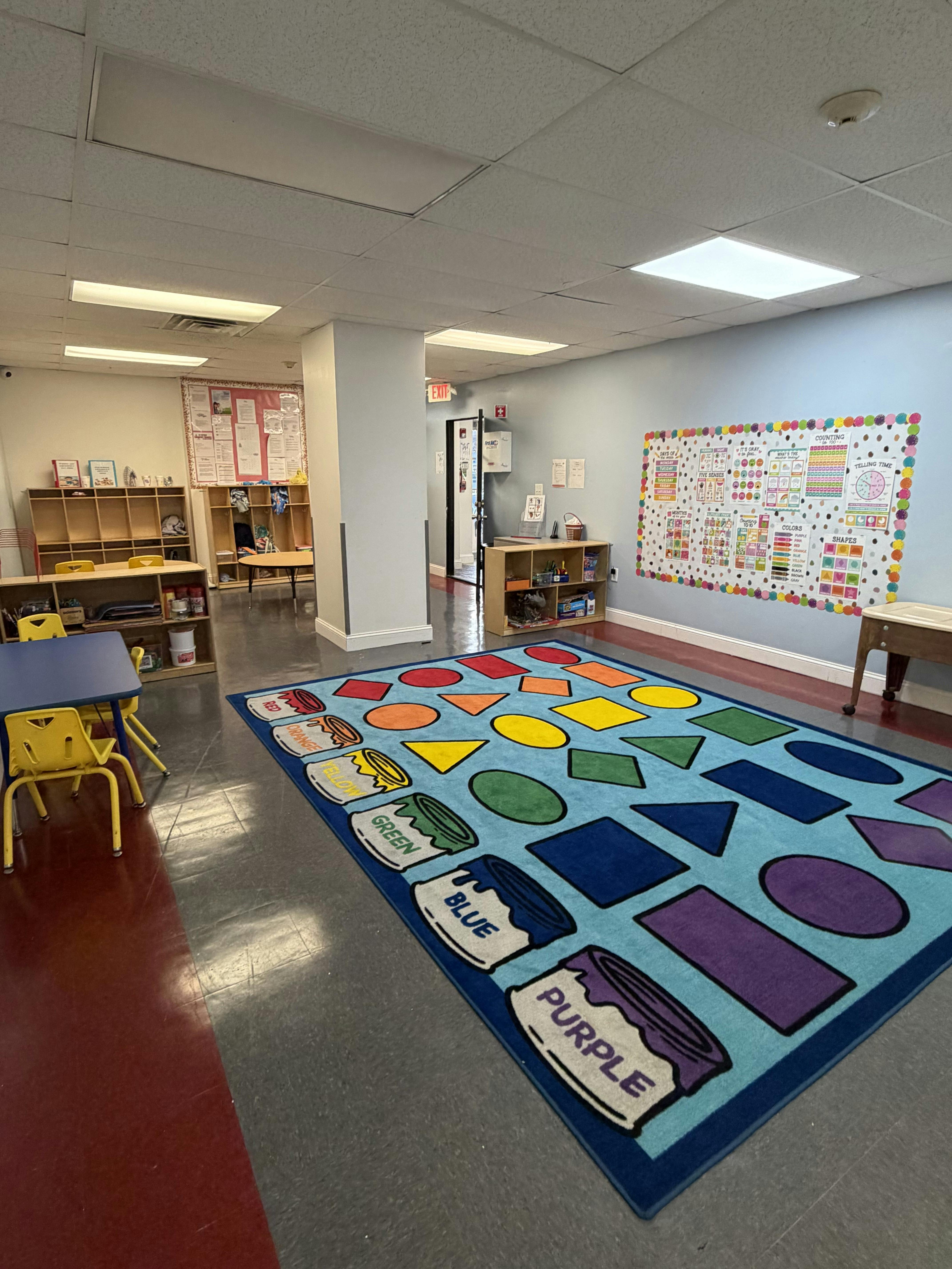Classroom with a colorful rug with shapes and paint cans, tables, and shelves