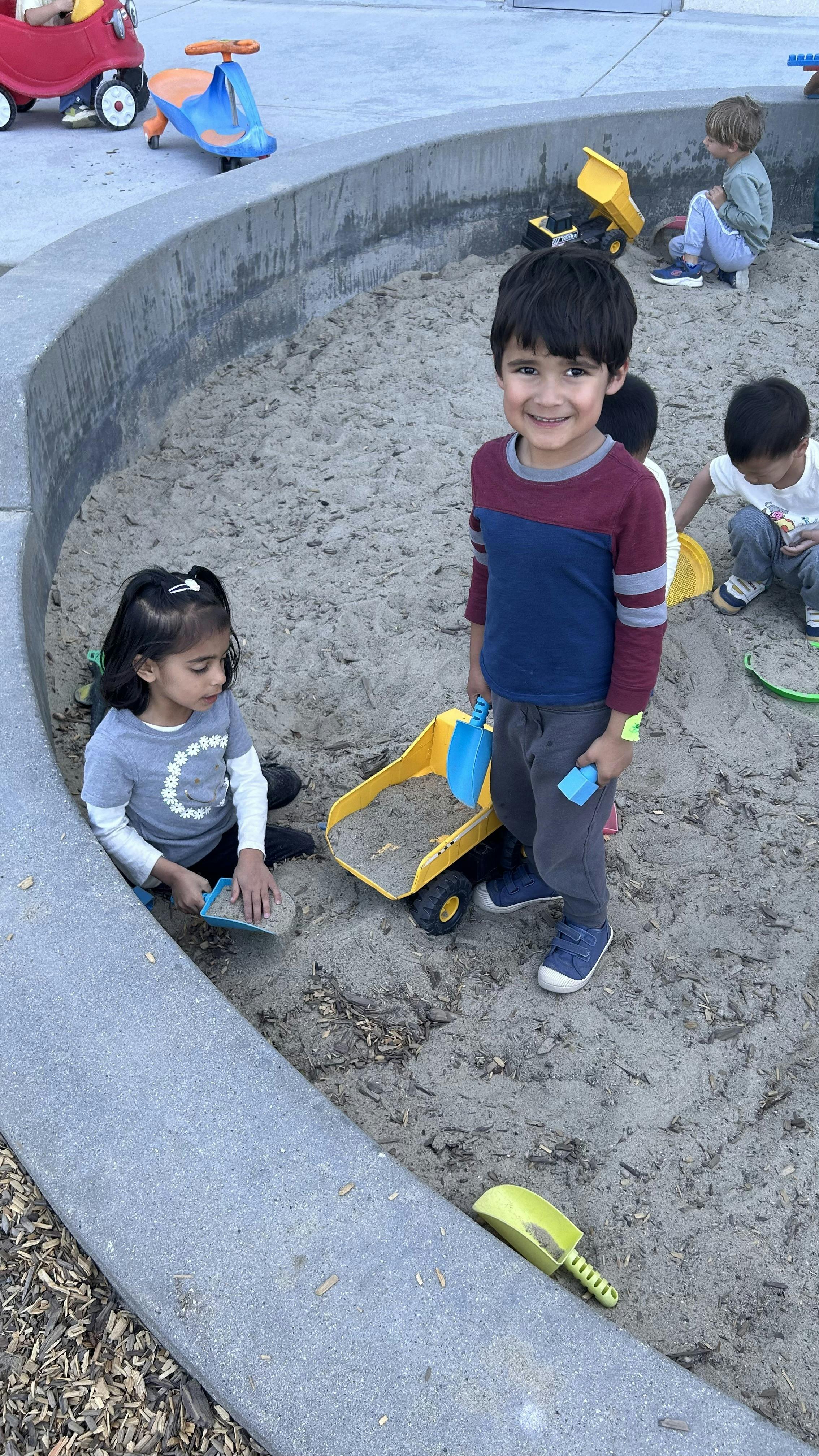 Children playing in a sandbox with toy trucks and shovels