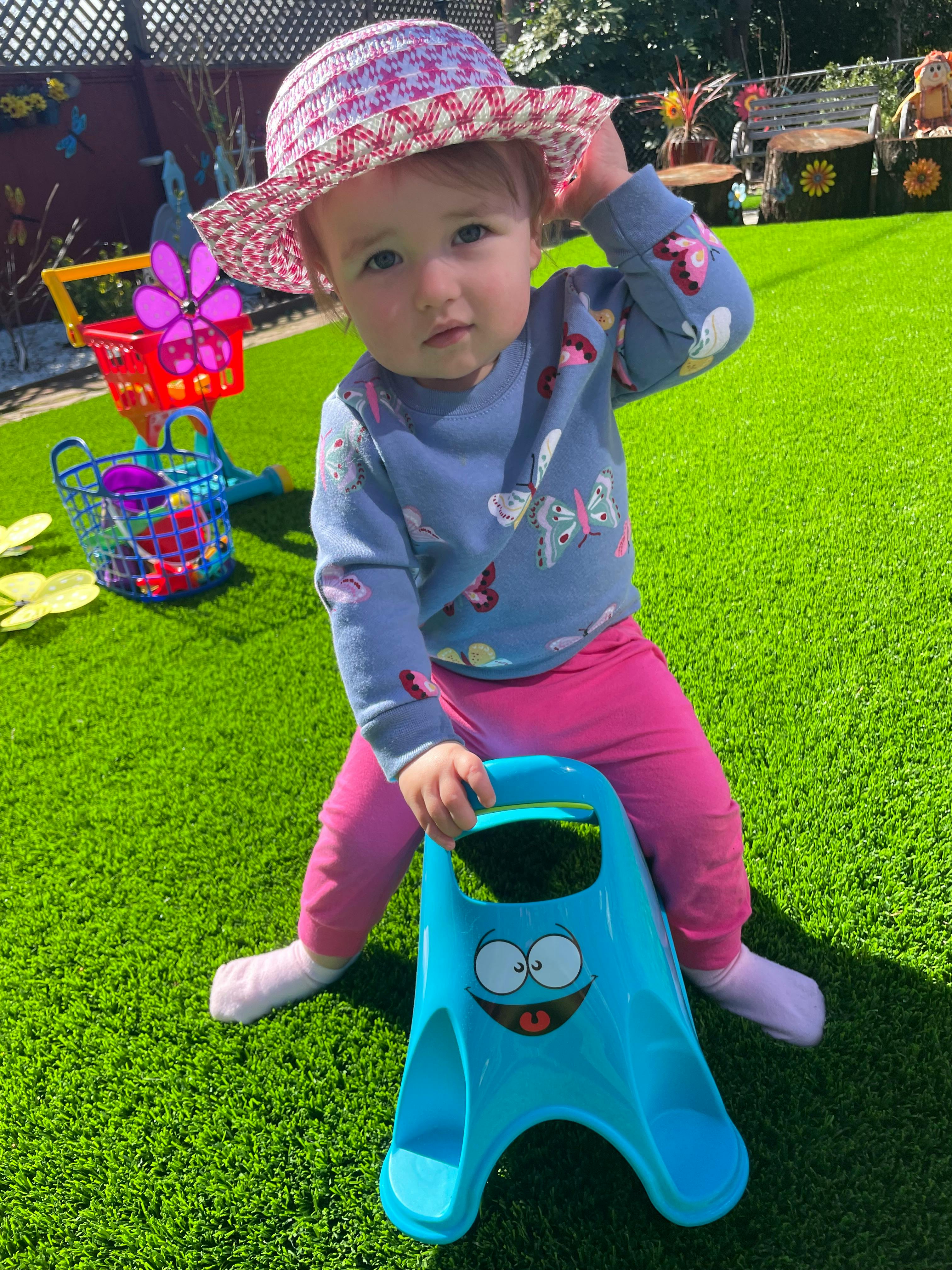 Child in a hat sitting on a blue toy on artificial grass