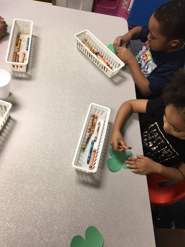 Two children coloring green paper cutouts at a table