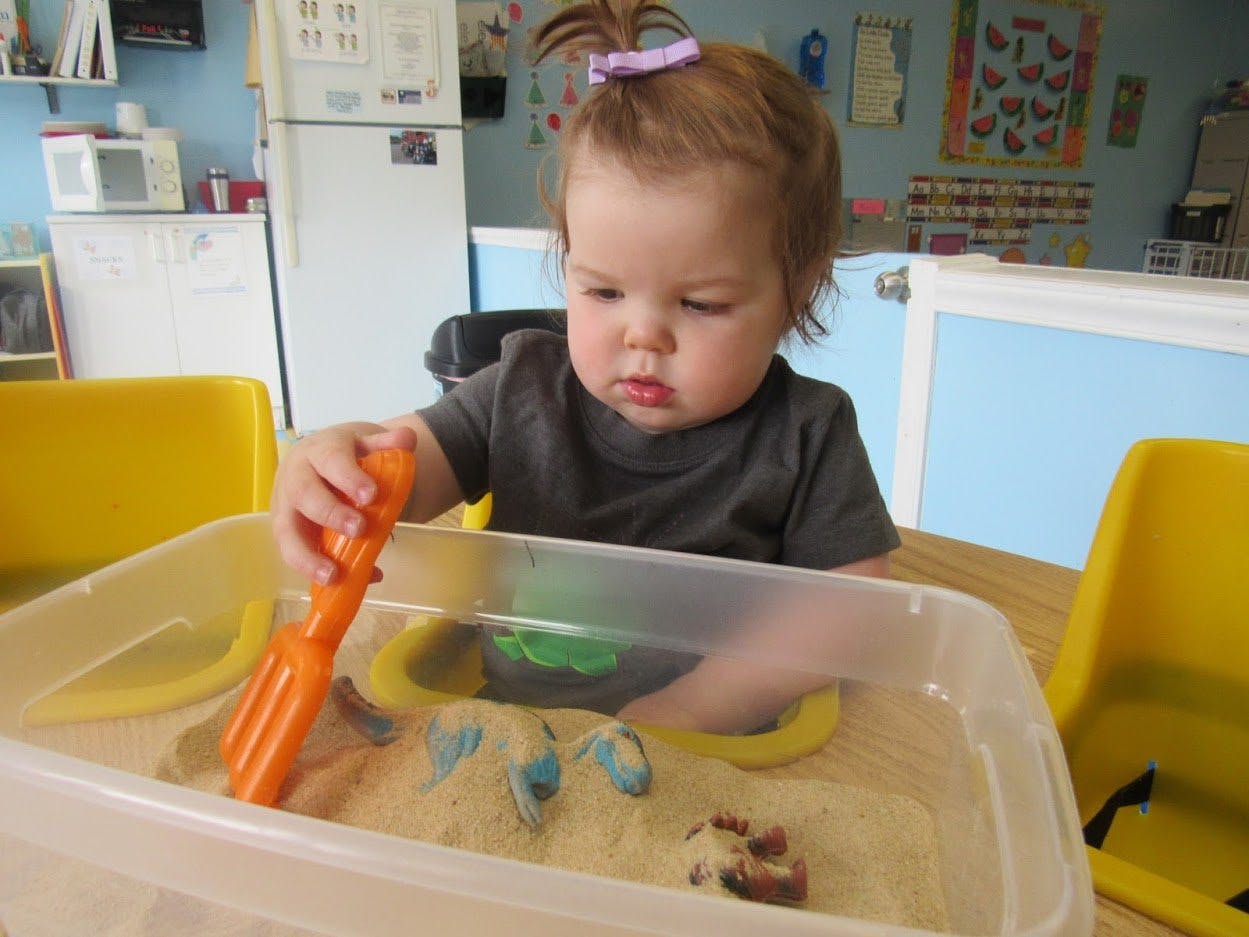 Child playing with sand and an orange toy in a clear bin