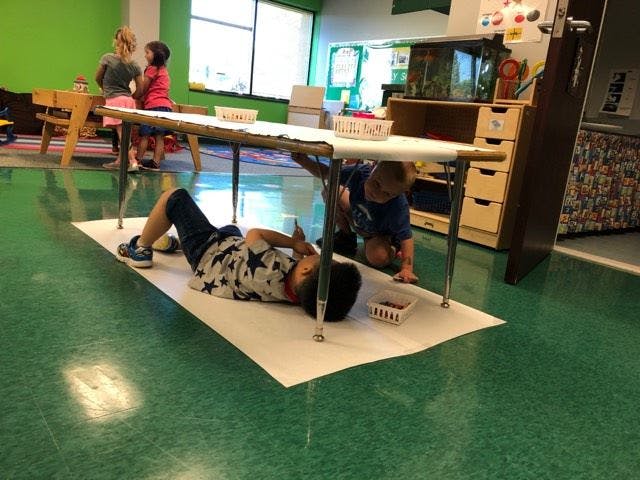 Child lying under a table while another child kneels beside them